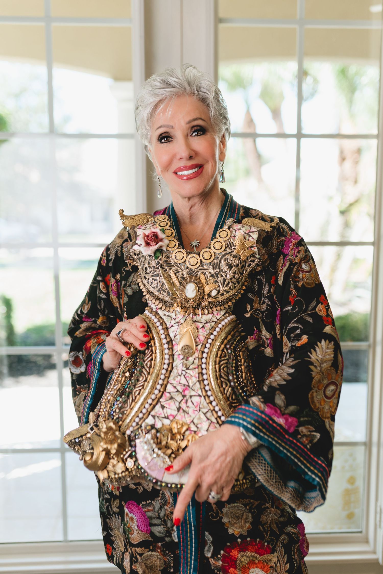 Elegant older woman with short gray hair holding ornate embroidered decor, wearing a floral kimono, smiling indoors.