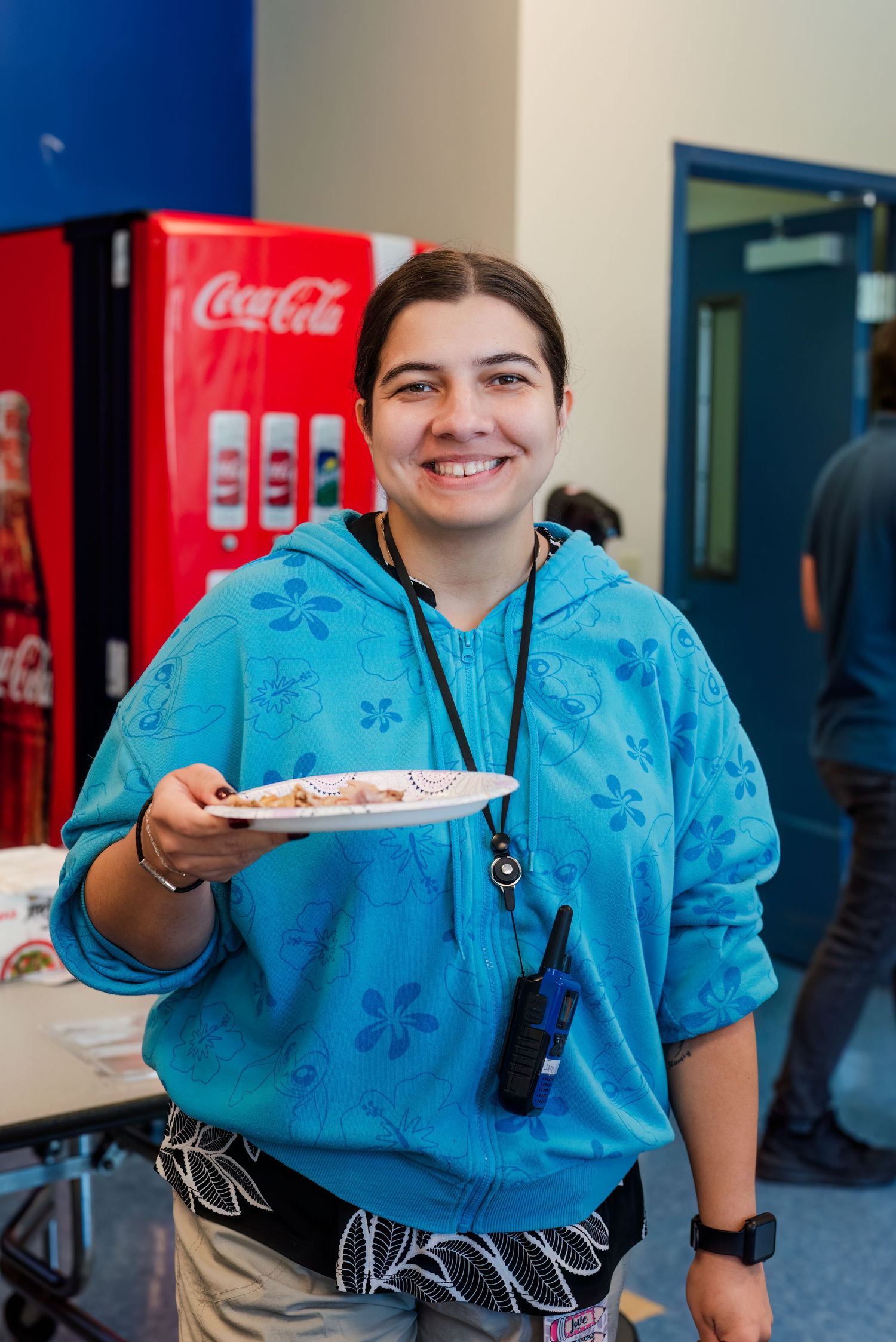 A person in a bright blue floral hoodie holds a plate while standing near a Coca-Cola vending machine.