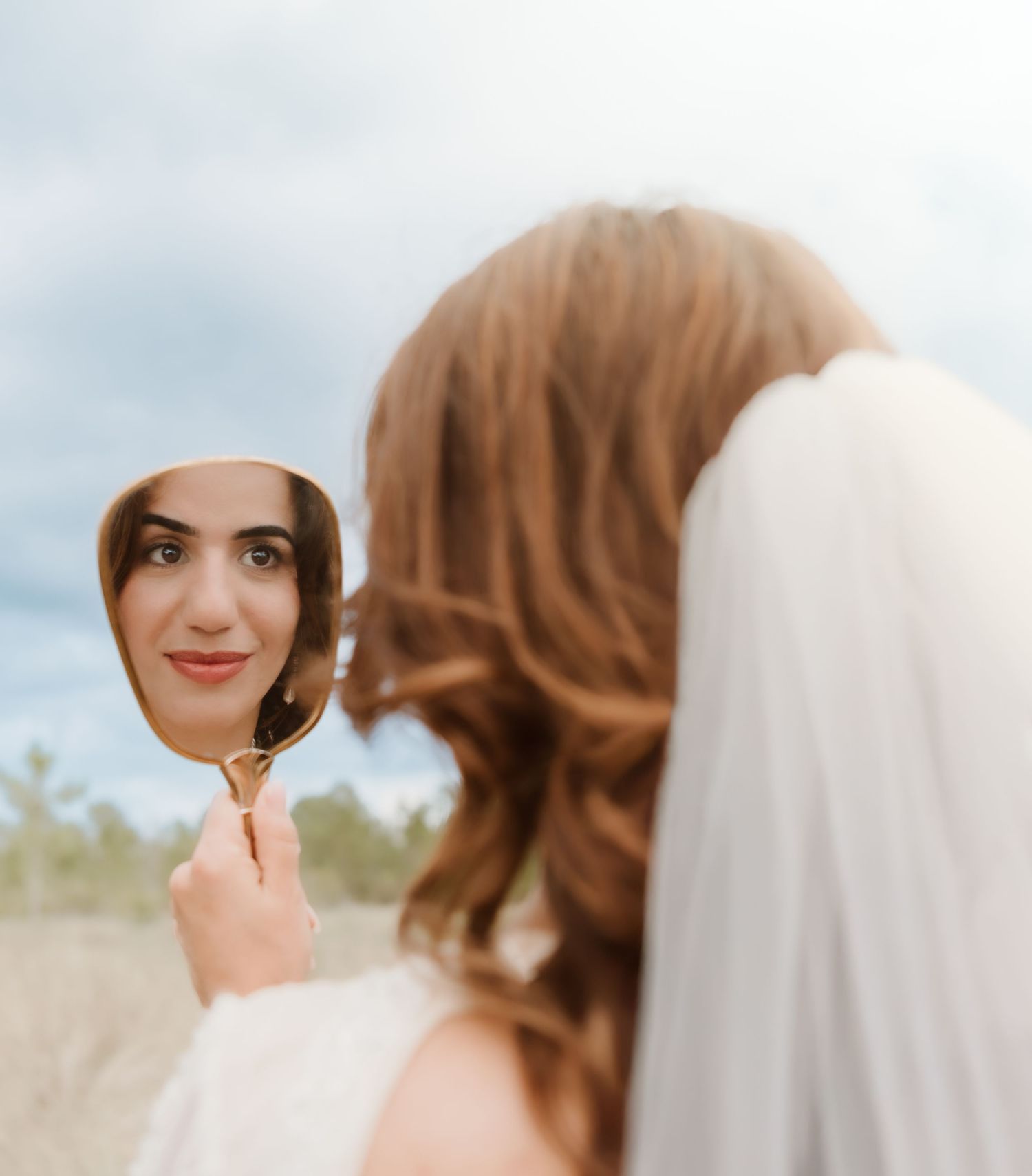 A bride holds up a handheld mirror reflecting her smiling face against a cloudy outdoor background.
