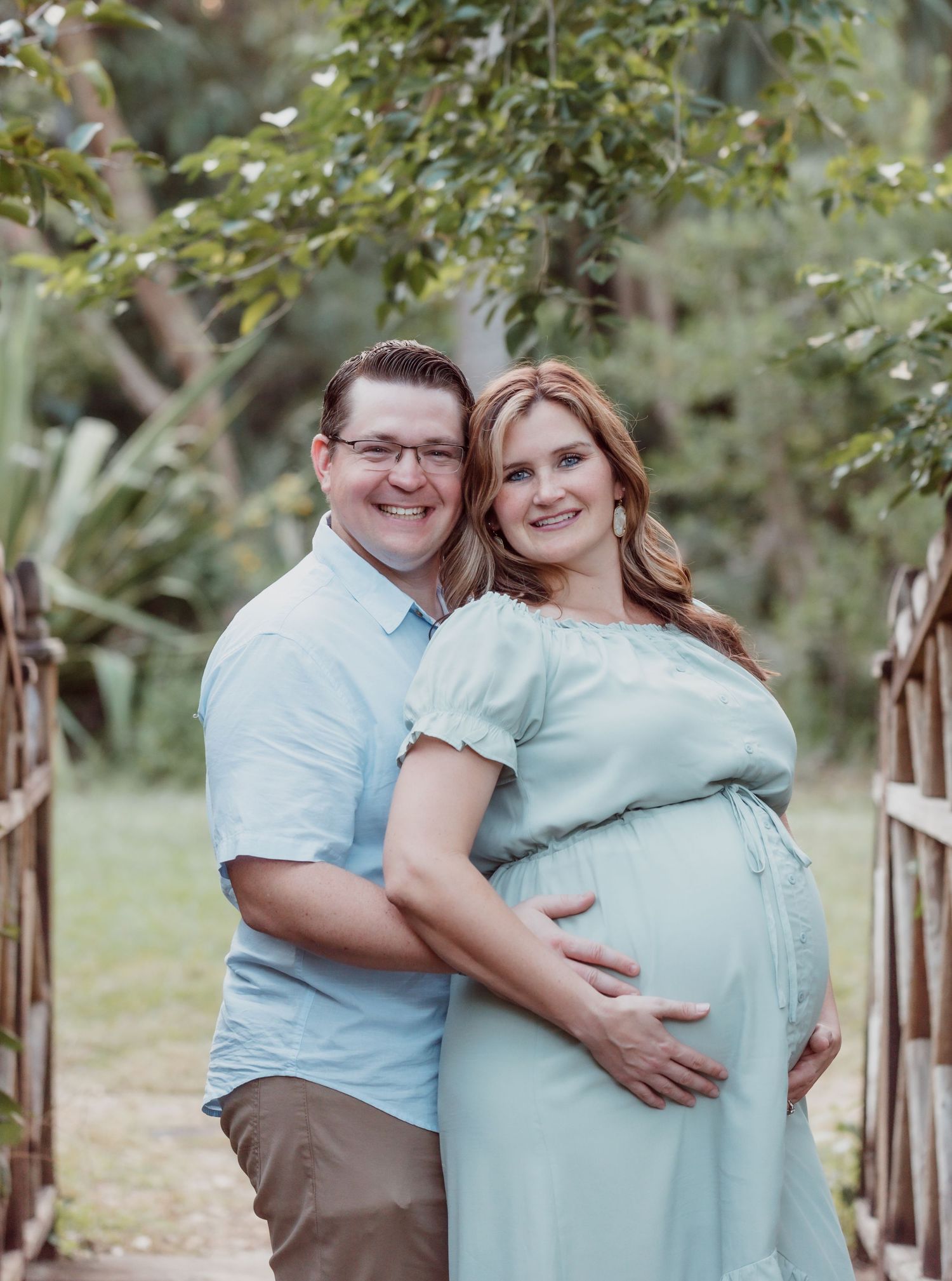 Couple poses for a maternity photo outdoors, wearing light blue and brown clothes while embracing under tree branches.