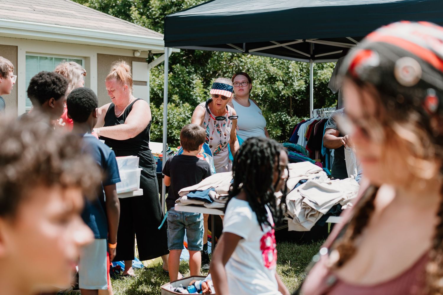 Large group gathered at an outdoor community event with white tent canopies and tables set up in a park setting.