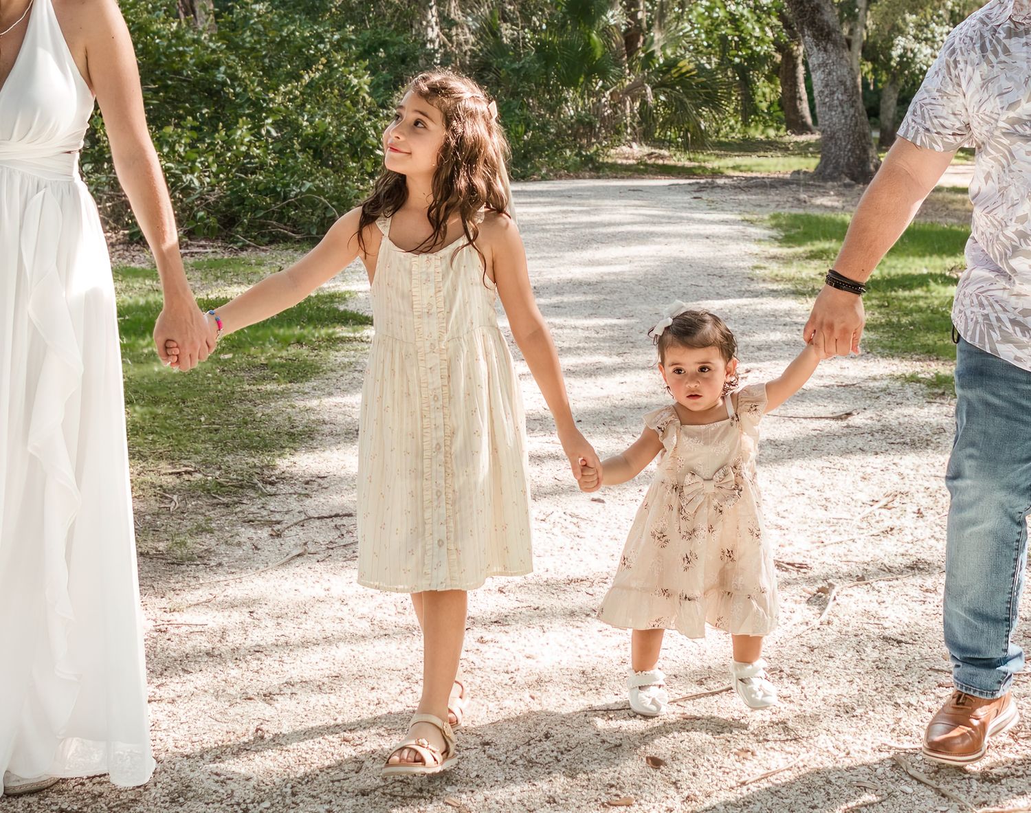 A group wearing neutral colored dresses and clothing walks together on a gravel path in a park setting.