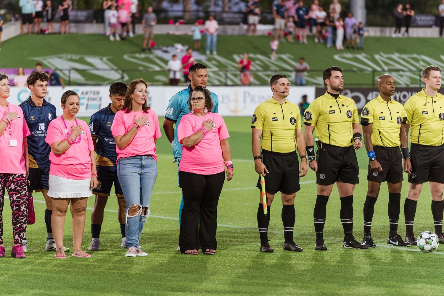 Soccer players and officials line up on a green field wearing pink and yellow uniforms during a pre-match ceremony.