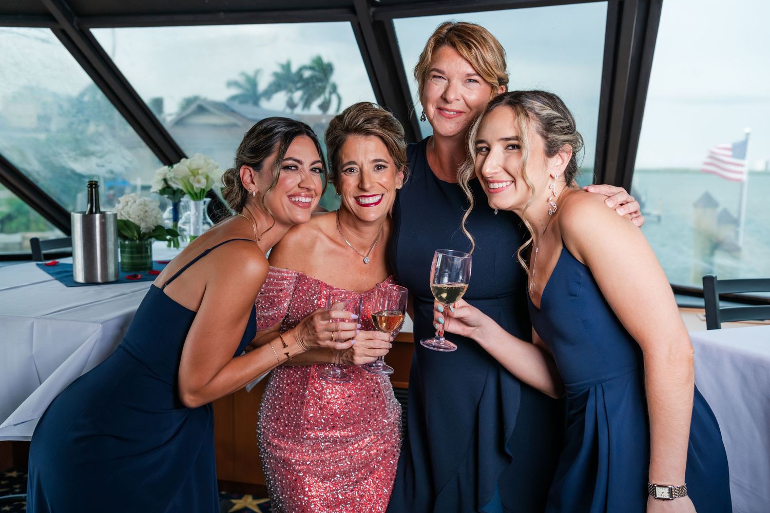 Four women in elegant attire smile and raise champagne glasses aboard a luxury yacht at a celebration.