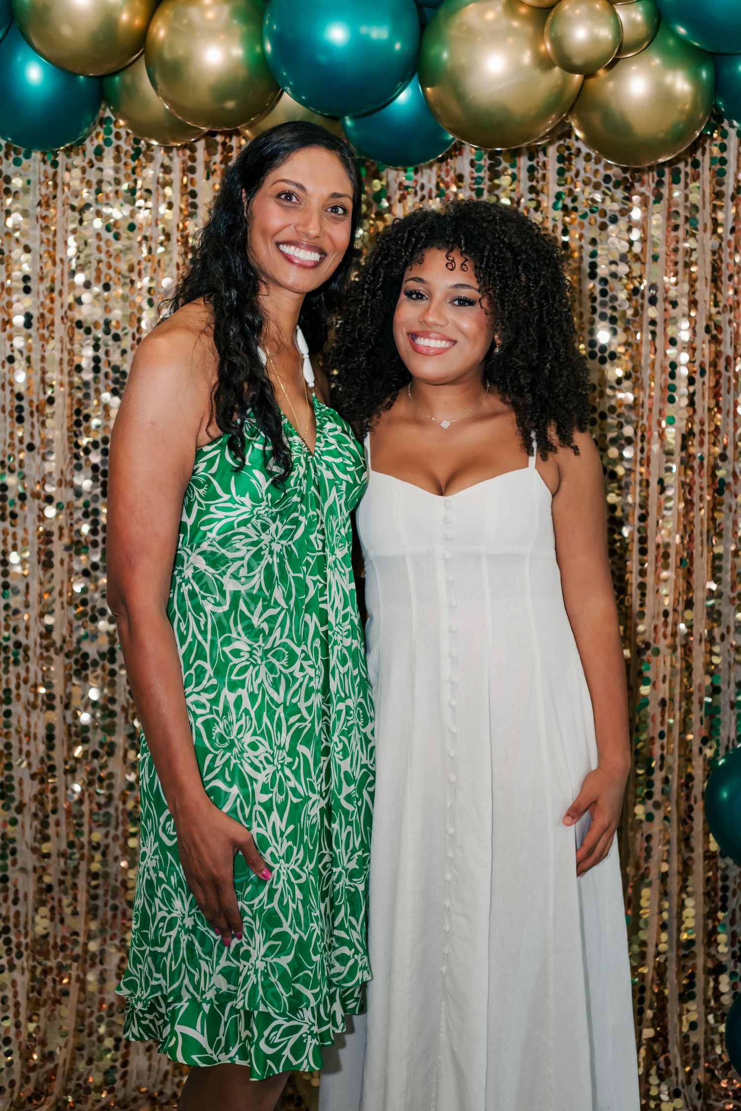 Two women smiling at a festive party with gold and teal balloon arch and sequin backdrop.