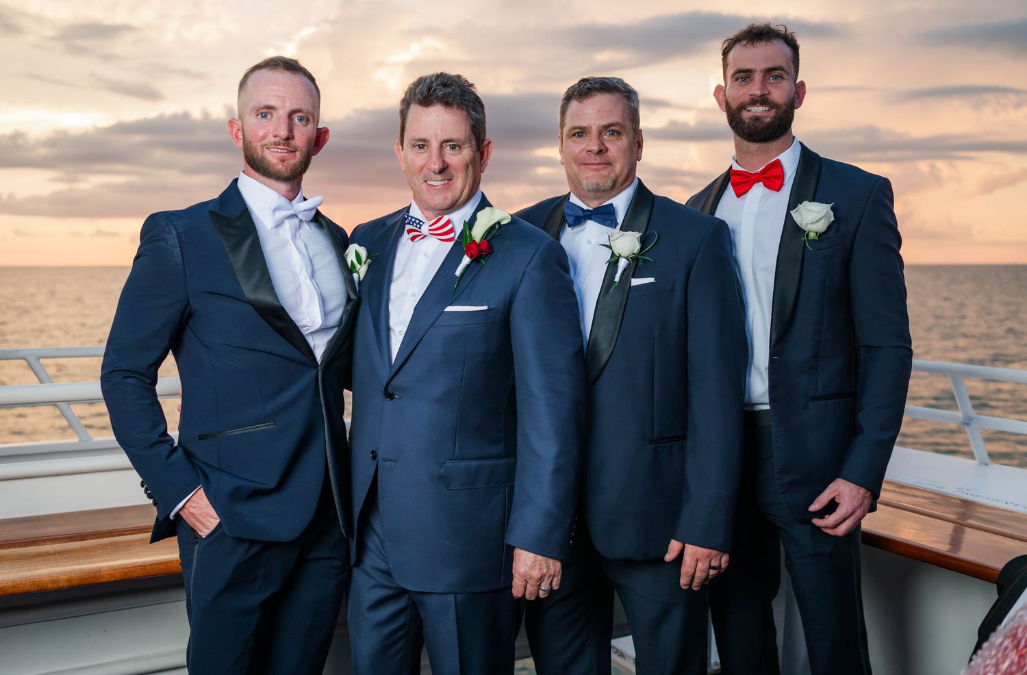 Four groomsmen in navy suits with floral boutonnieres pose on a cruise ship deck at sunset.