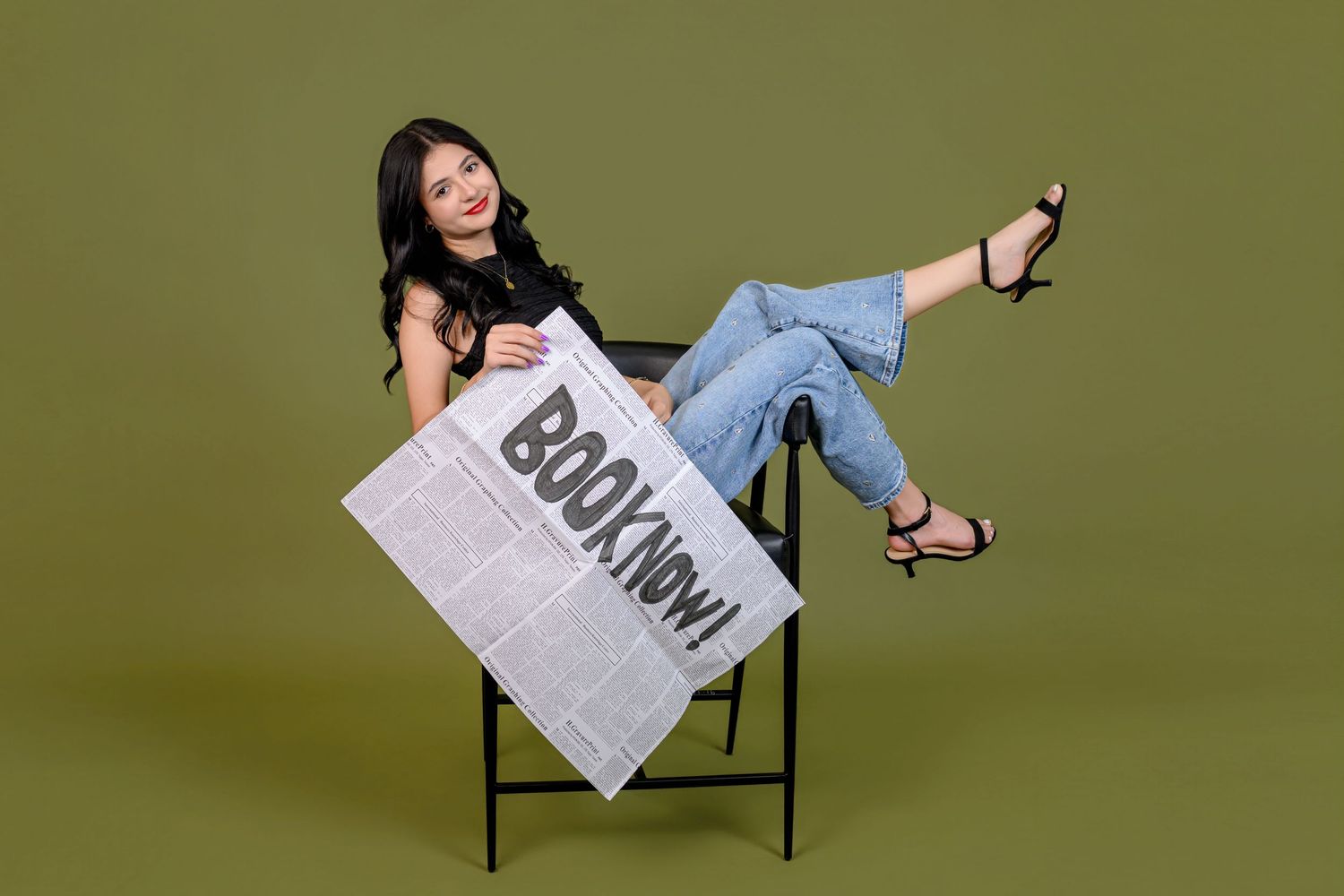 Smiling woman in jeans holding a 'Book Now!' sign while sitting on a chair against an olive green background.