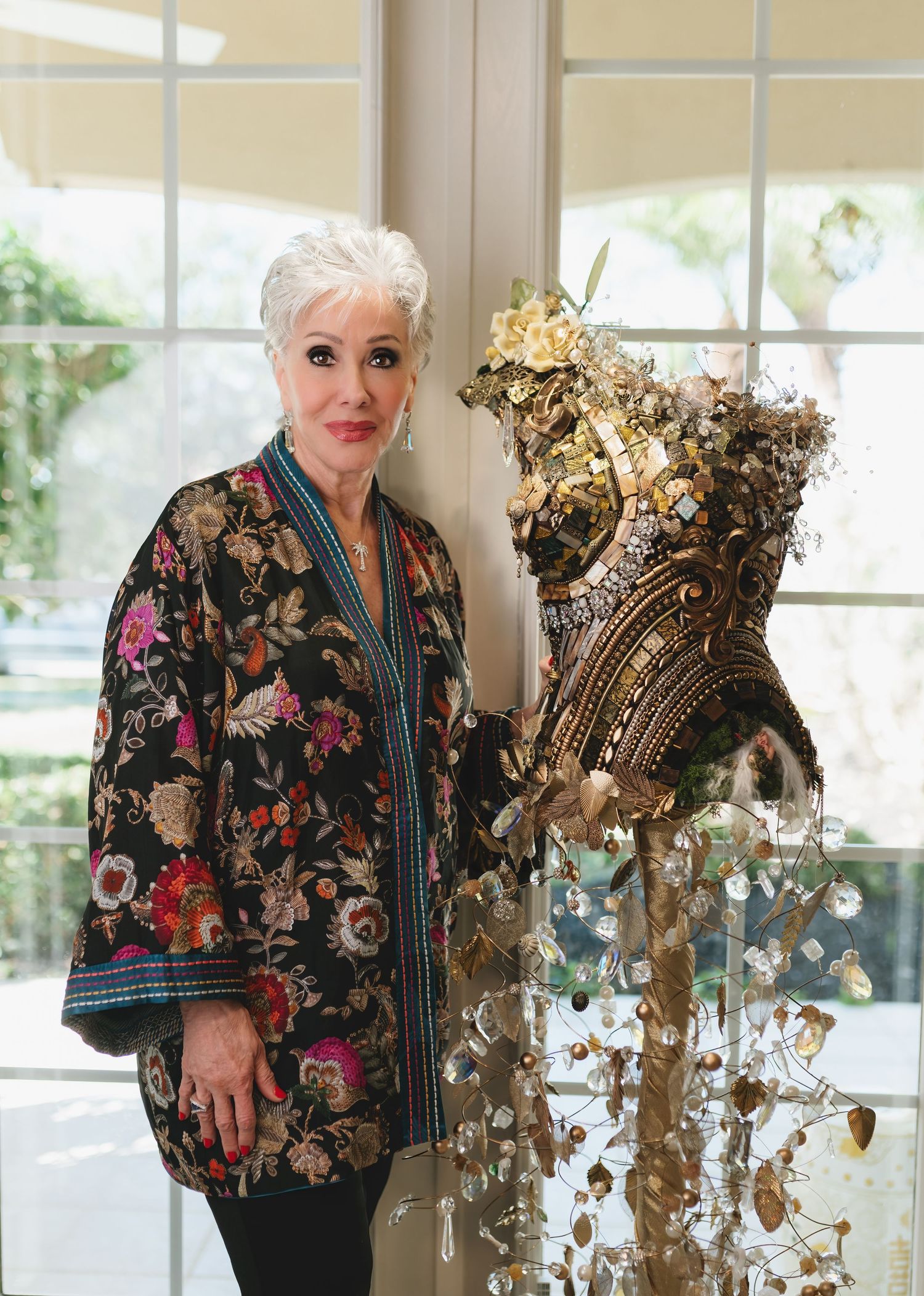 Elegant woman in floral jacket stands beside an ornate gold mosaic corset sculpture adorned with flowers indoors.