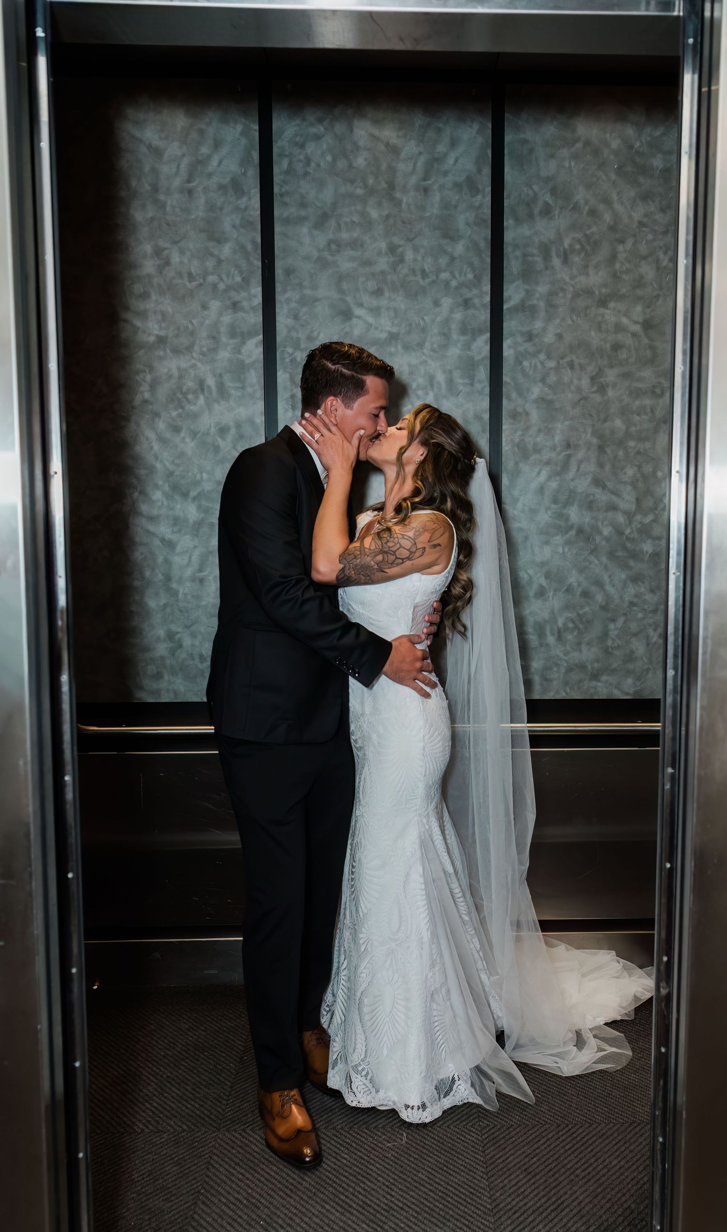Bride and groom sharing a romantic kiss inside an elevator, bride wearing a lace mermaid gown with veil.