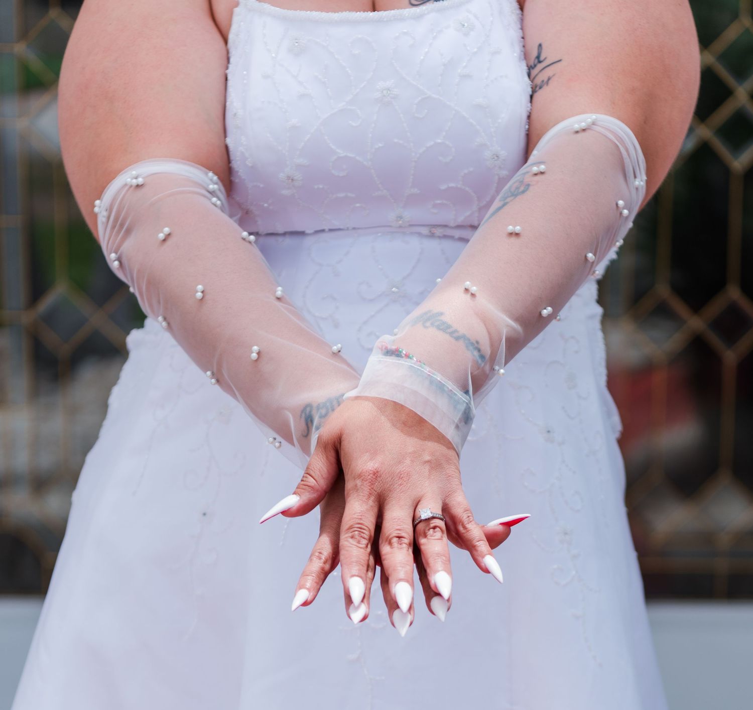 Bride wearing sheer tulle gloves and white wedding dress displays long white acrylic nails with red polish tips.