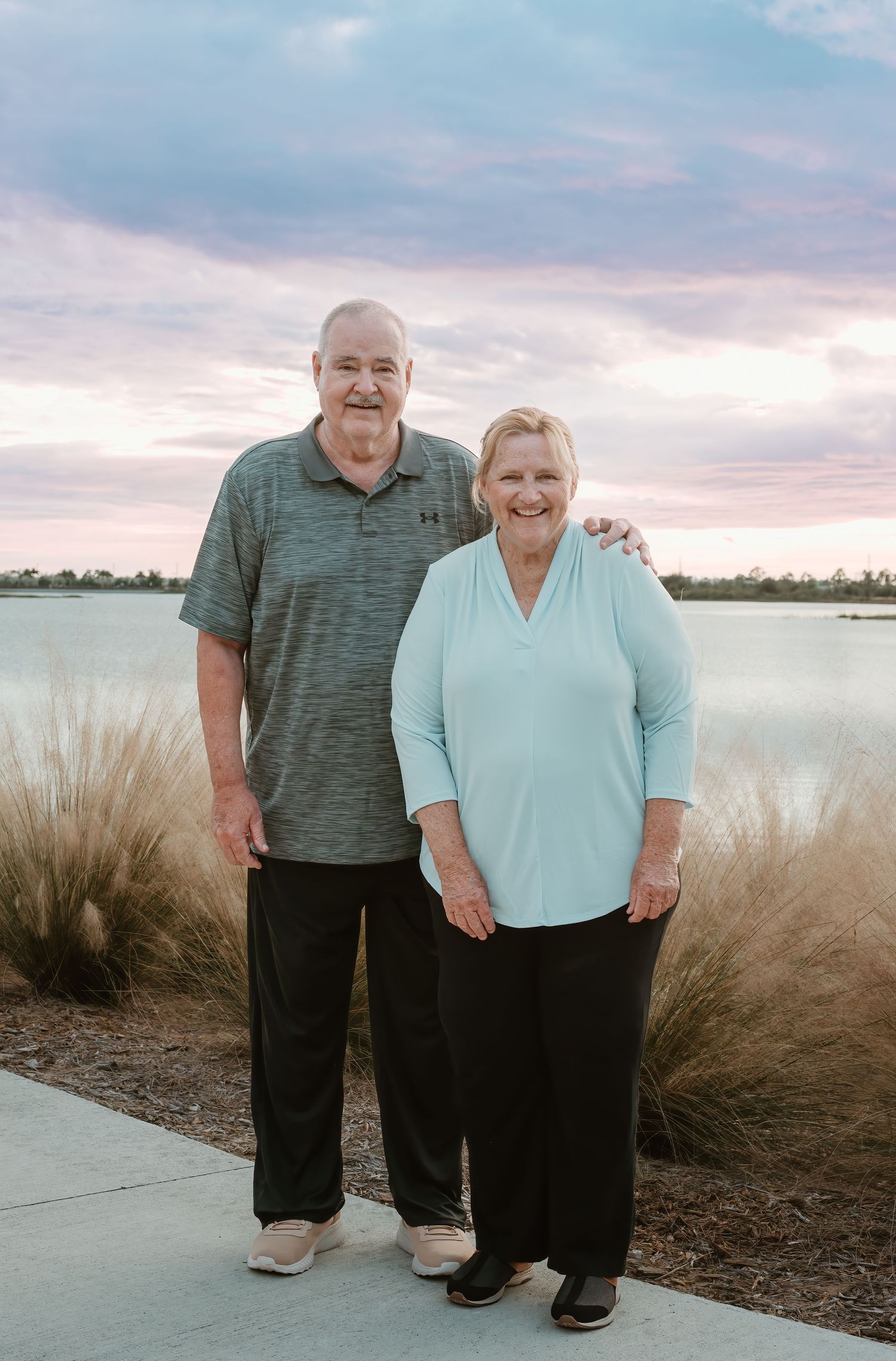 A content couple stands together on a lakeside path at sunset, with ornamental grasses in the foreground.