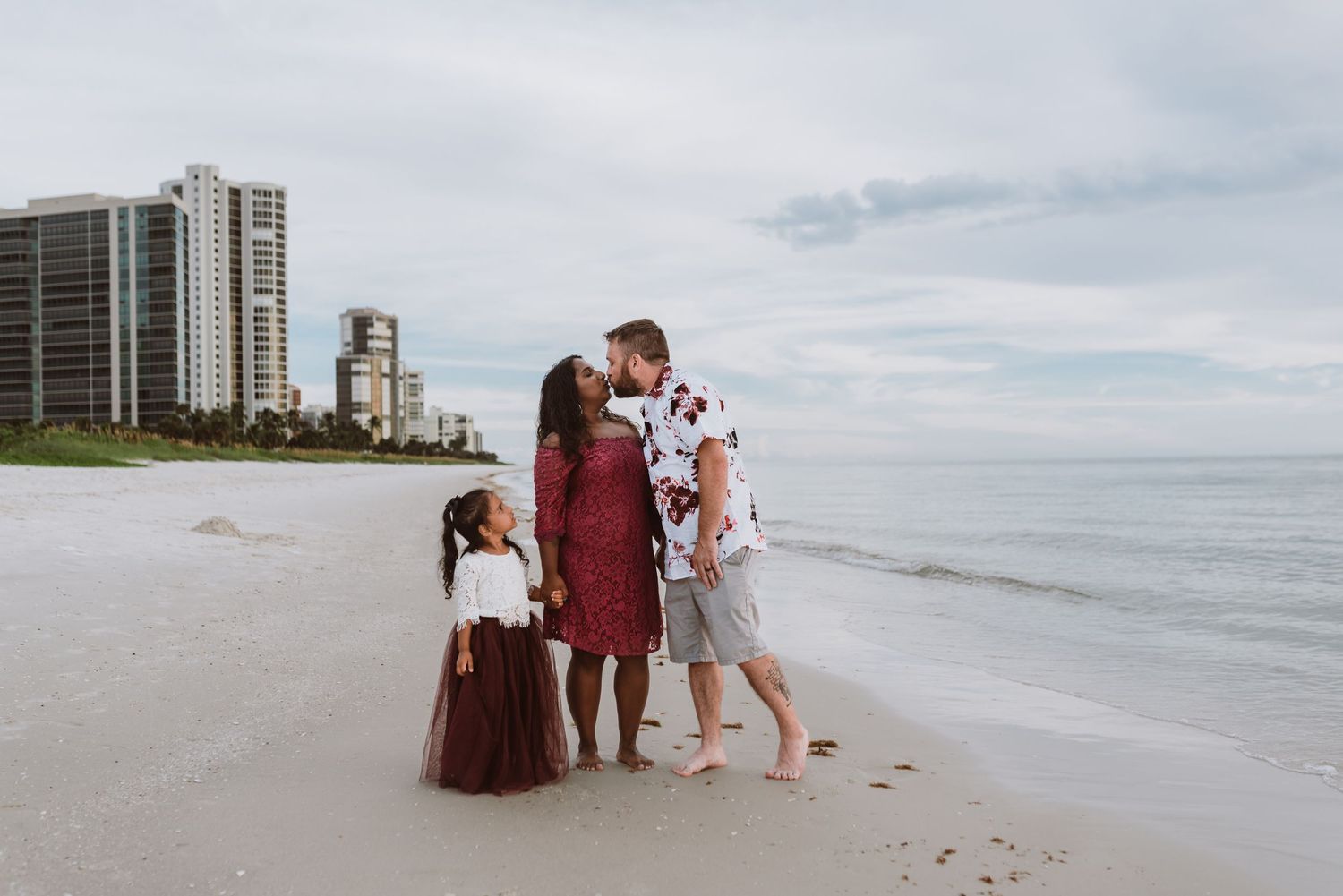 A small group stands together on a sandy beach with high-rise buildings visible in the background on a cloudy day.