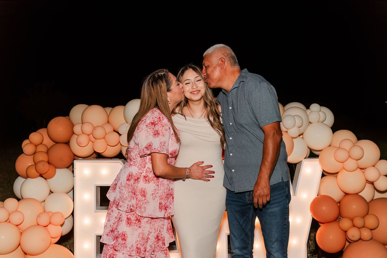 Three people pose in front of a peach and white balloon arch display at a celebratory event.