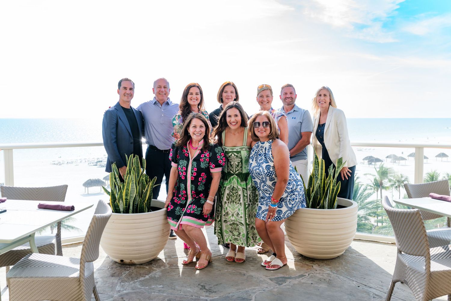 Group of ten people posing together on a beachfront terrace with ocean views and tropical plants.