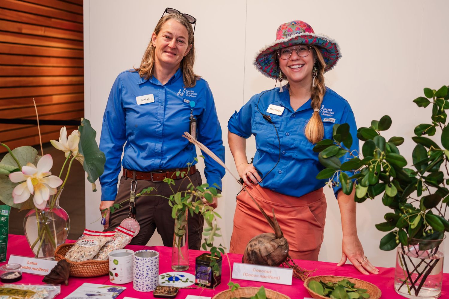 Two women in blue shirts smile at a plant display table with tropical specimens at an indoor gardening event.