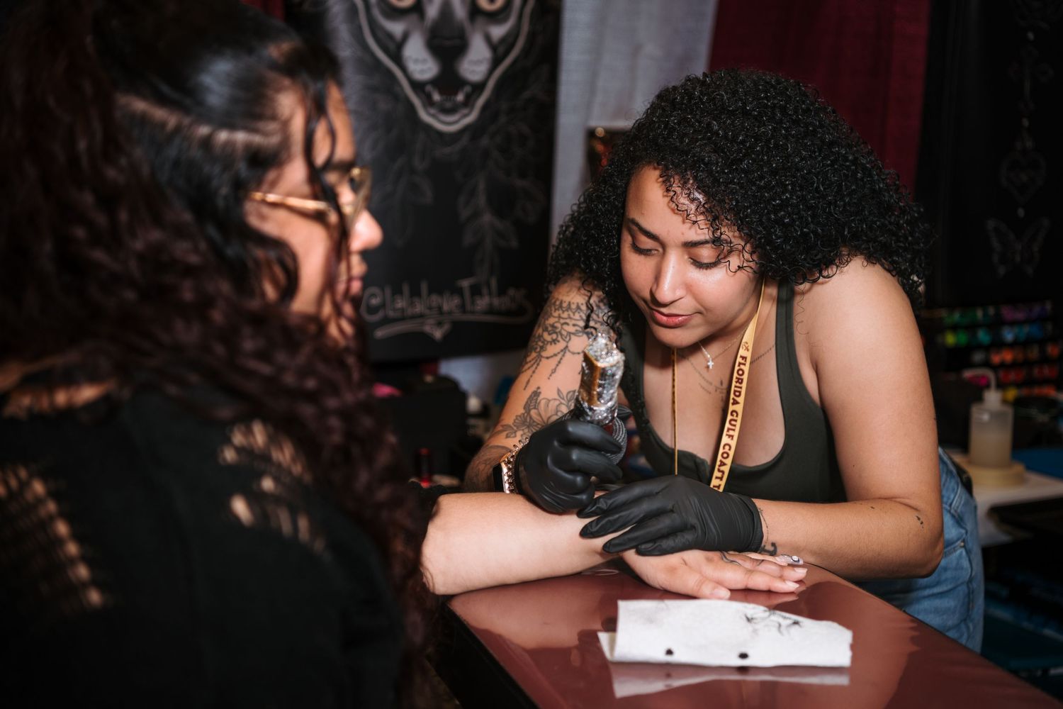 Female tattoo artist wearing black gloves tattooing a clients arm at a tattoo studio.