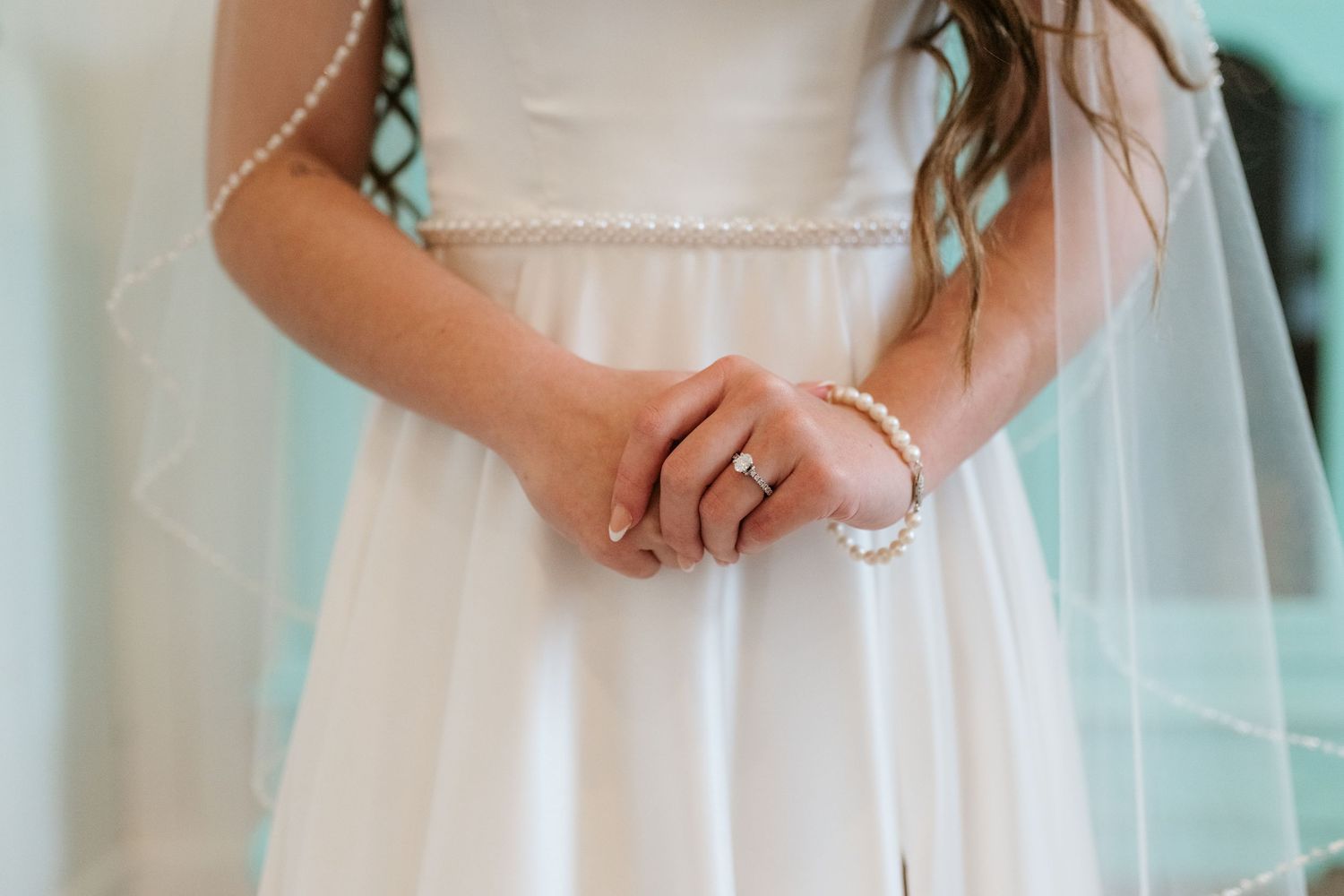 Bride in white wedding dress with pearl bracelet and engagement ring clasping hands at waist with veil detail.
