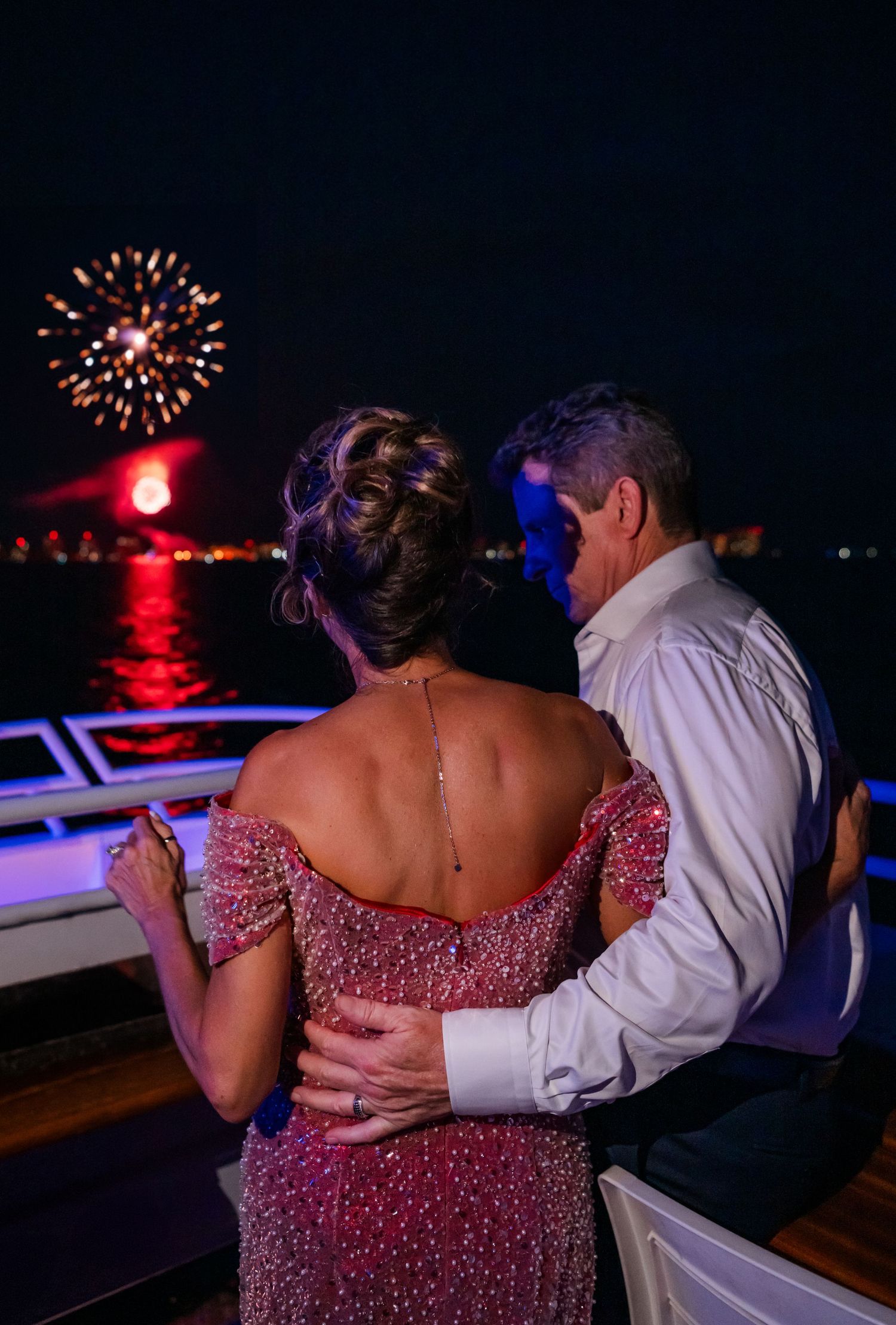 Couple embracing on a yacht at night watching colorful fireworks reflecting over the water, woman in elegant beaded gown.