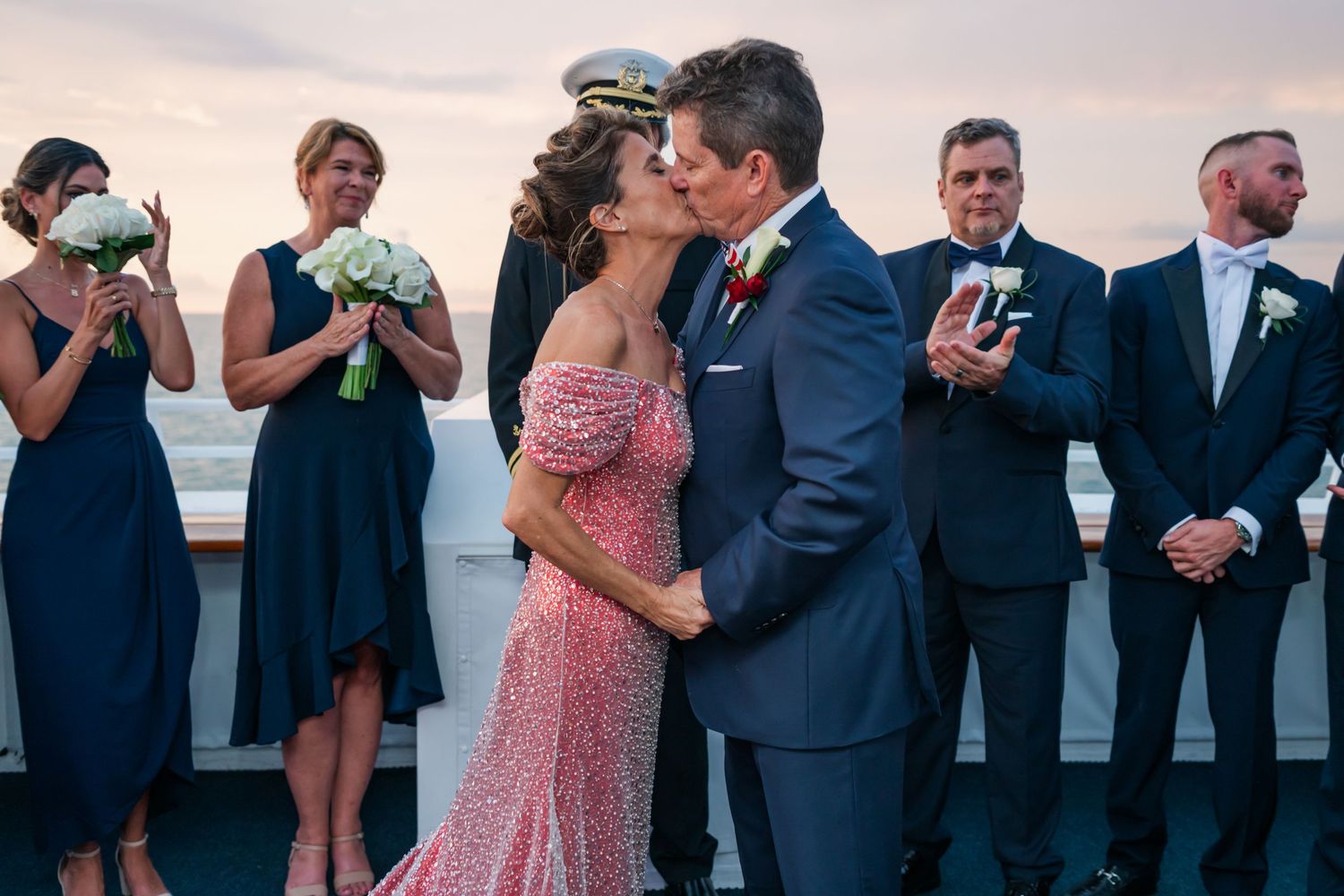 Couple shares first kiss at outdoor wedding ceremony on a ship deck with bridesmaids in navy and groomsmen behind them.