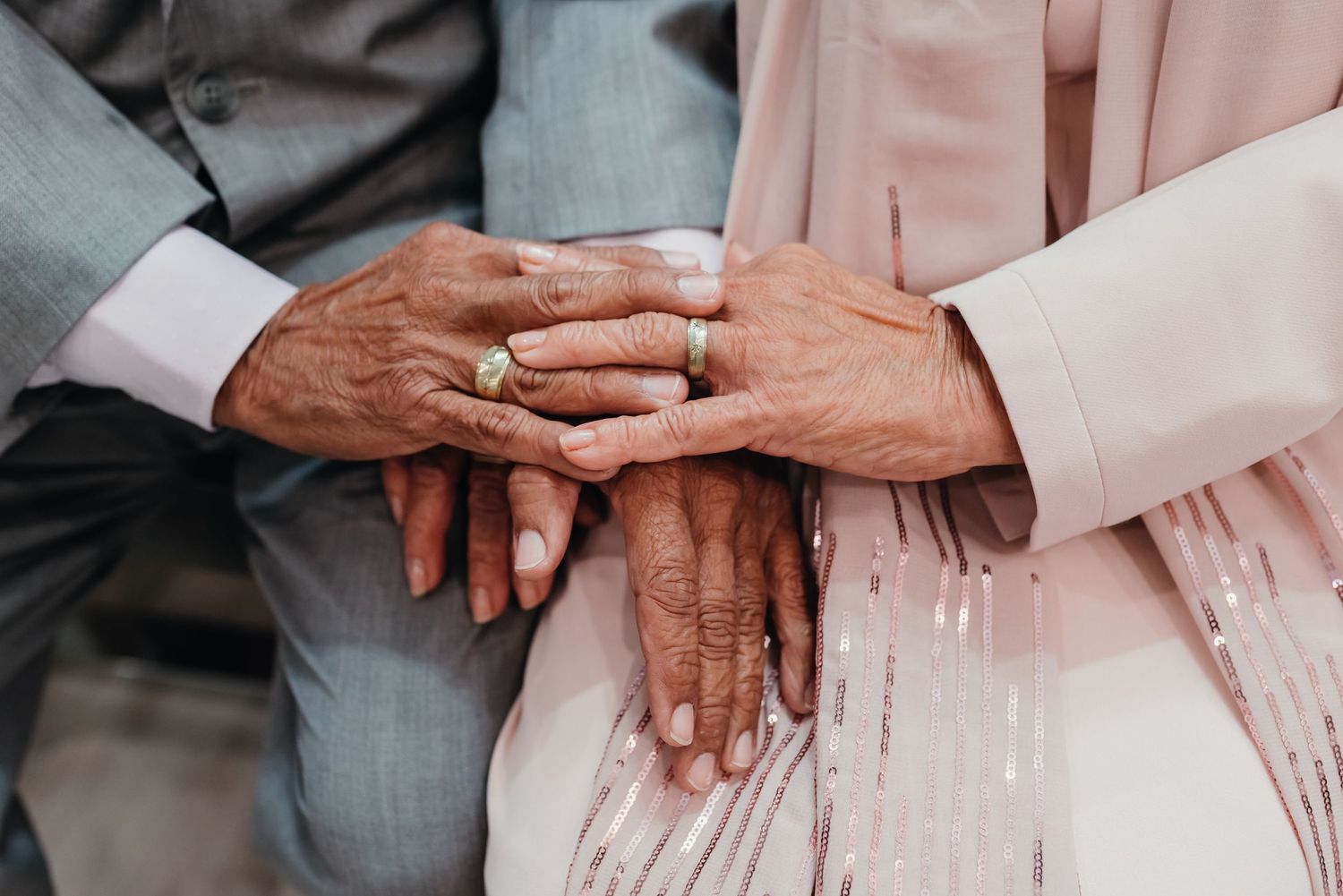 Close-up of hands with wedding rings touching tenderly in elegant gray and pink formal attire.