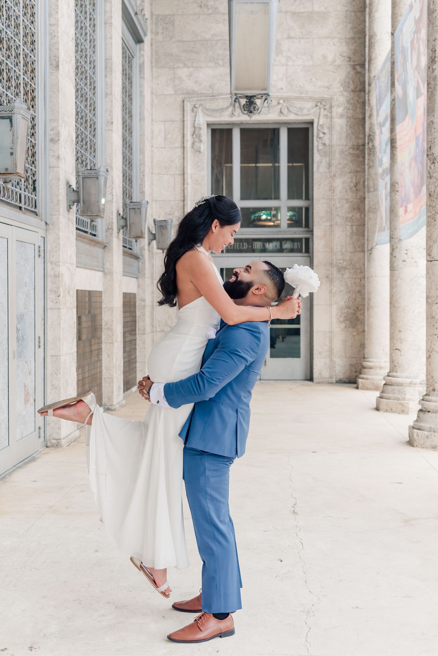 A couple shares a romantic moment in formal attire against an ornate marble building with columns.