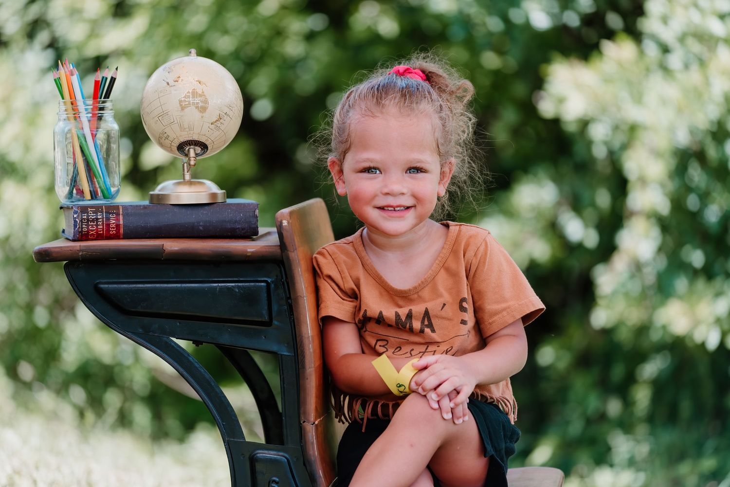 A young child sits at a classic wooden desk with globe and pencils outdoors in a sunny garden setting.
