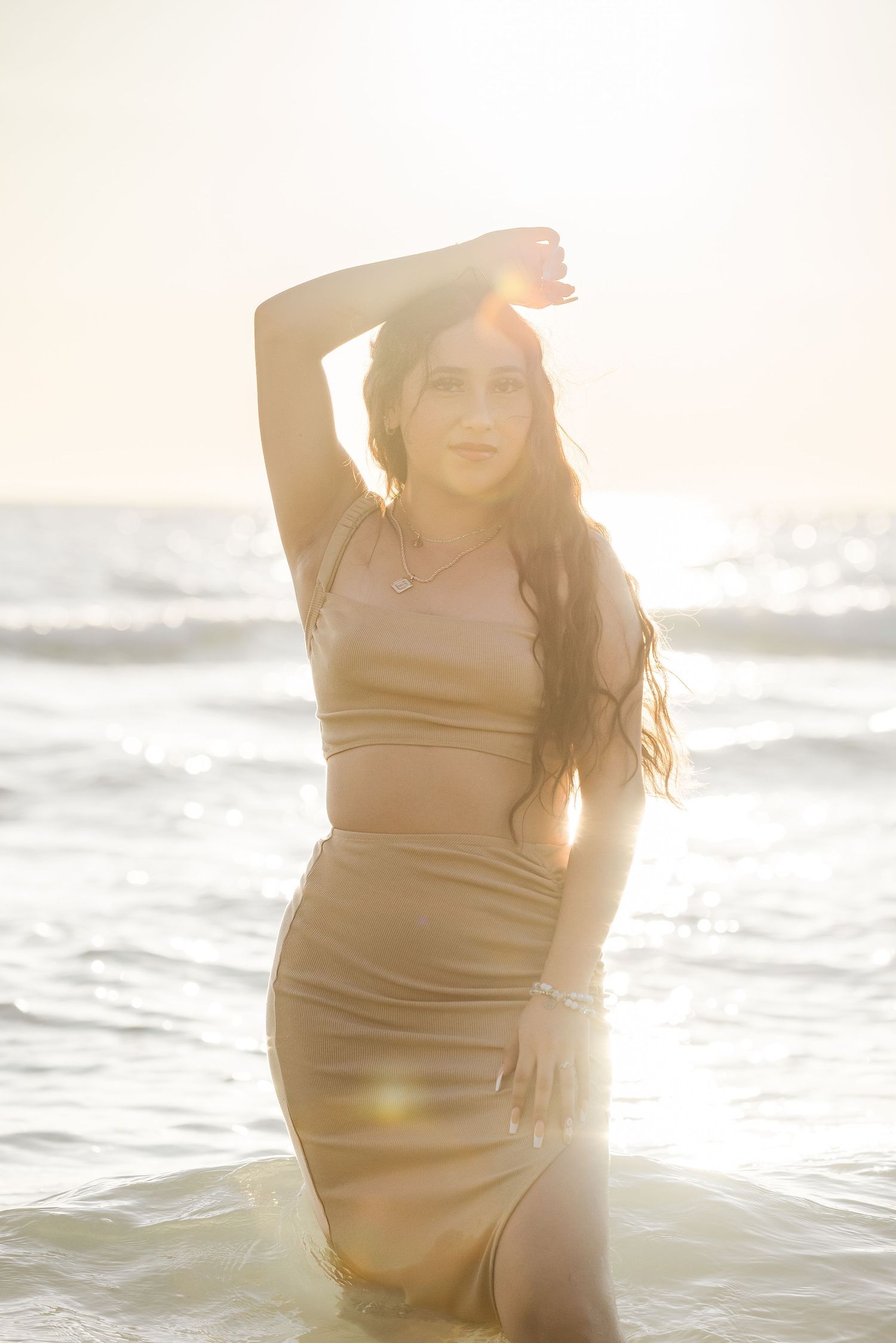 Person in a light colored dress poses at the beach during golden hour with sun flare and ocean waves.