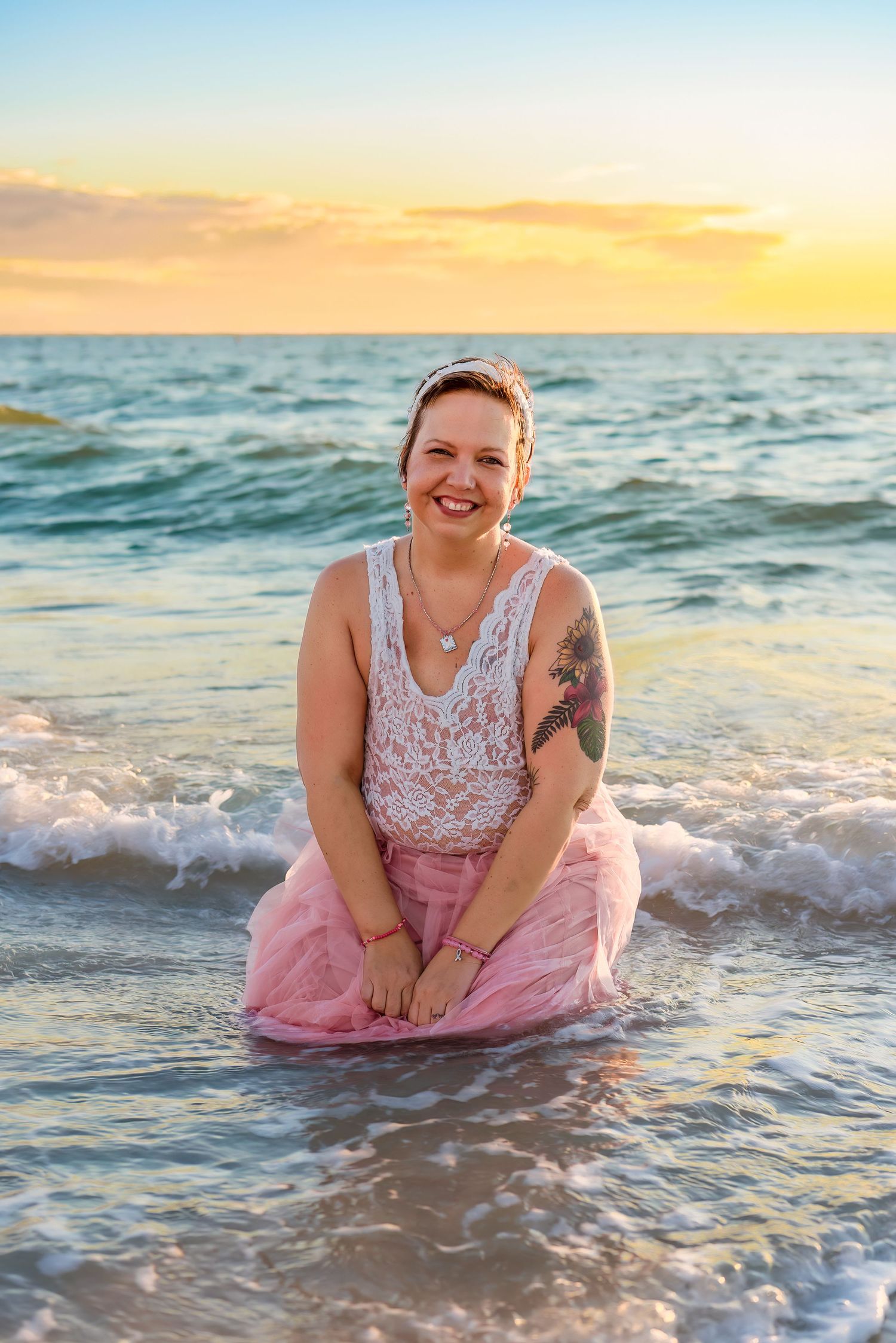 Someone in a pink tulle skirt and lace top poses playfully in the ocean waves at sunset.