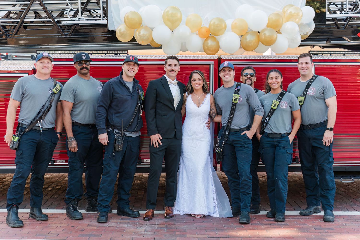 Wedding couple poses with firefighters in uniform in front of a decorated red fire truck with gold and white balloons.