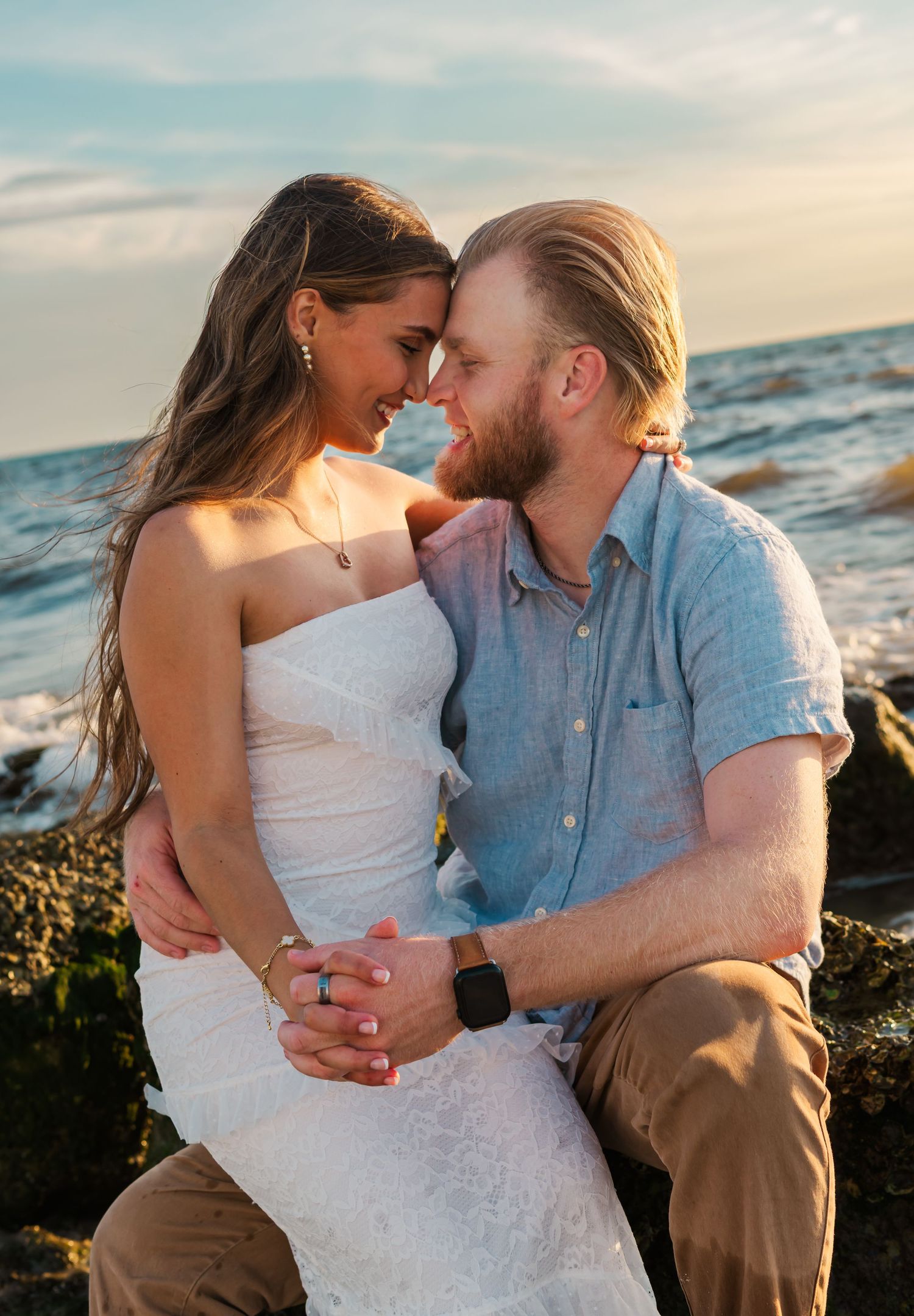 Couple sharing a romantic moment on rocky shoreline at sunset, woman in white dress, man in blue shirt.