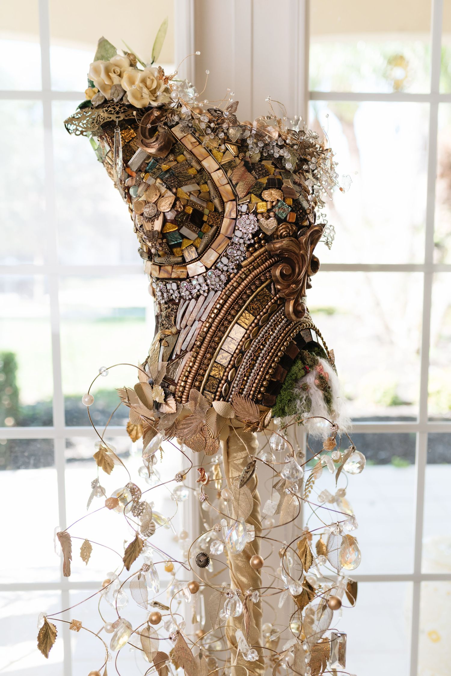 Intricate mosaic and jeweled corset dress form sculpture displayed near a bright window with cascading crystal accents.