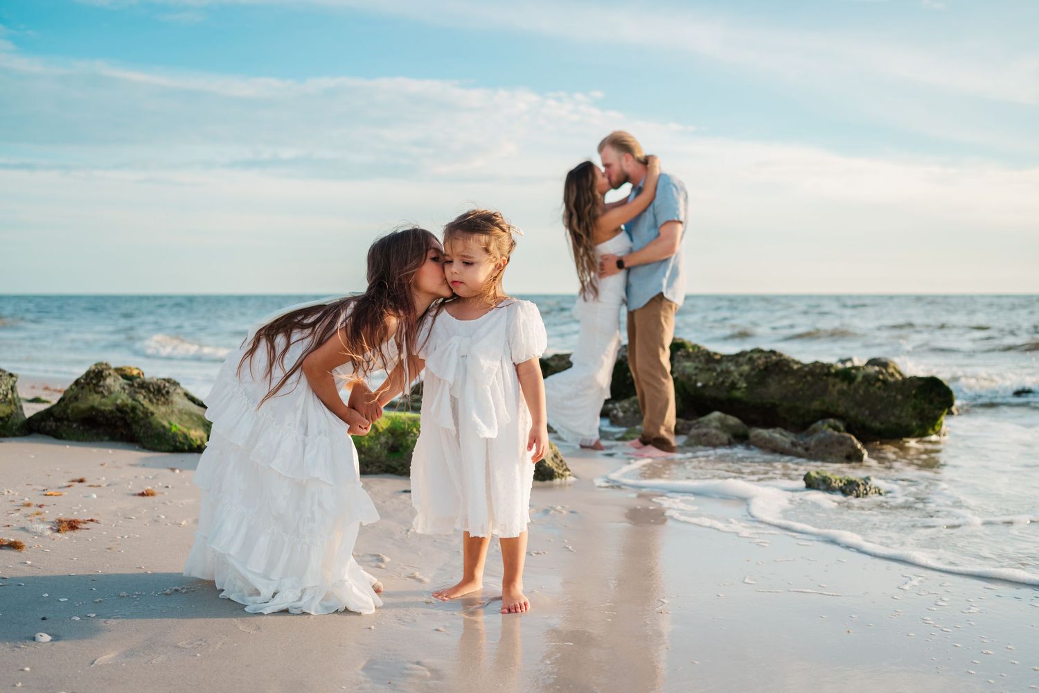 Family beach photo with couple kissing by rocks while two girls in white dresses play near the ocean shore.