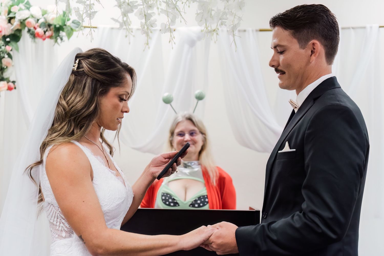 Bride and groom exchange vows at wedding ceremony with officiant wearing alien antennae headband in background.
