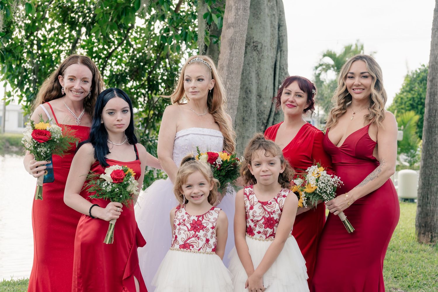 A group in red dresses and formal attire poses together for wedding party photos in an outdoor garden setting.