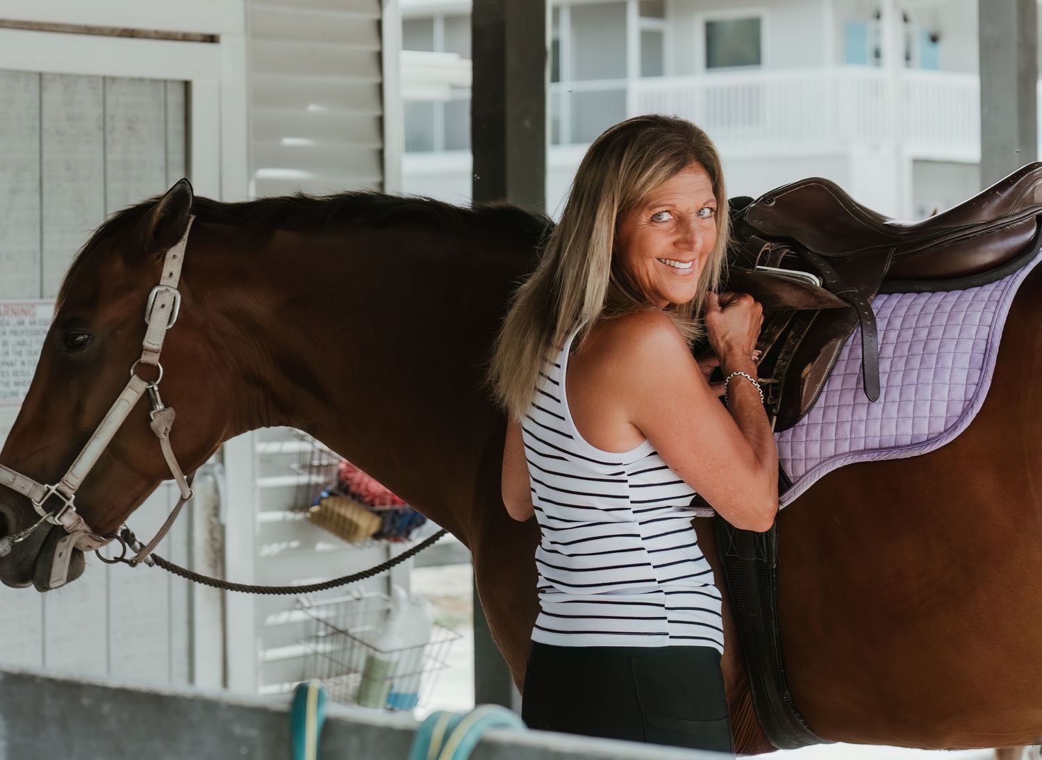 Someone in a striped tank top interacting with a bay horse in a barn setting.