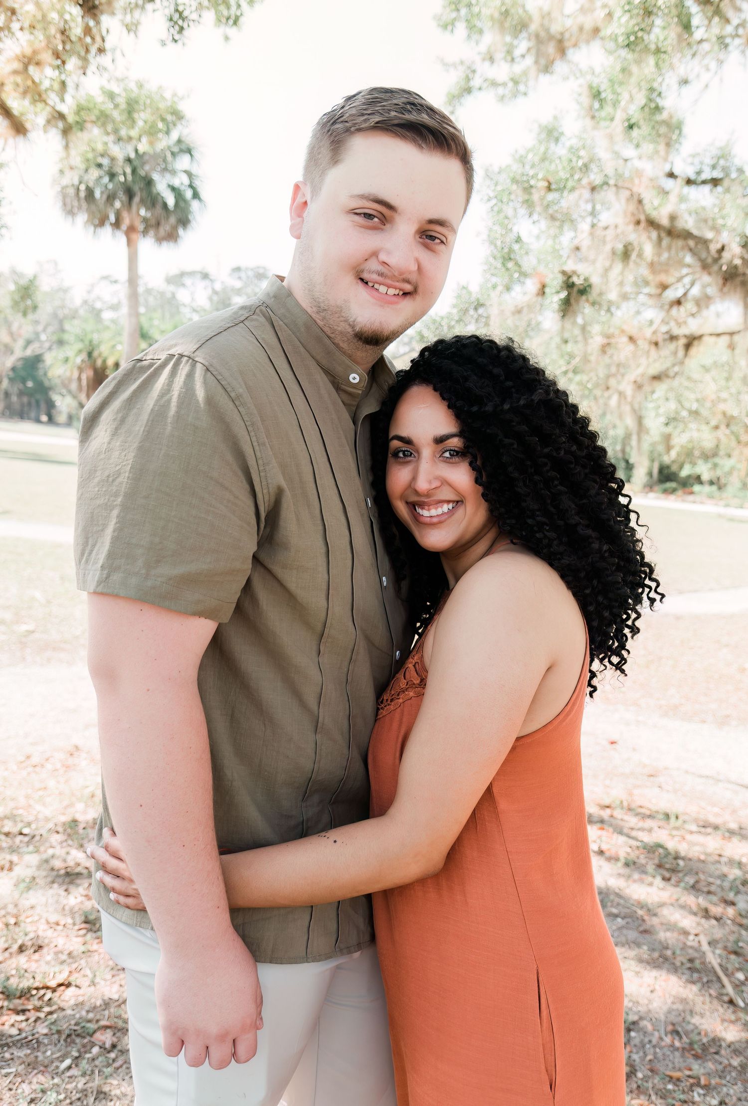 A couple embraces outdoors in a natural setting with palm trees and dappled sunlight in the background.