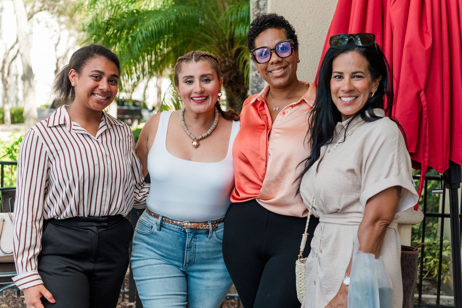 A group of people smile and pose together on a patio with red umbrellas in the background.