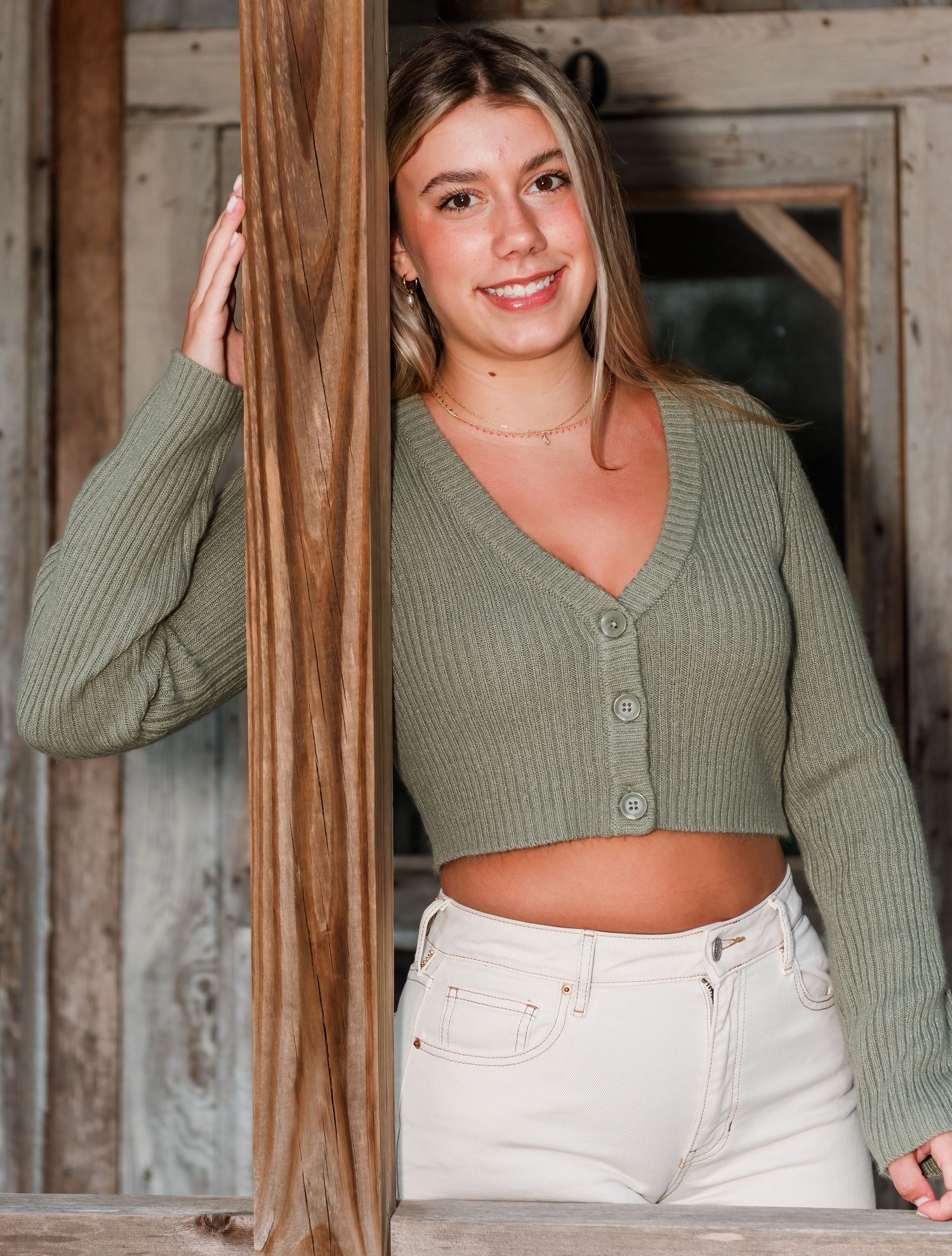 Person wearing sage green cropped cardigan and white jeans poses against rustic wooden door frame.