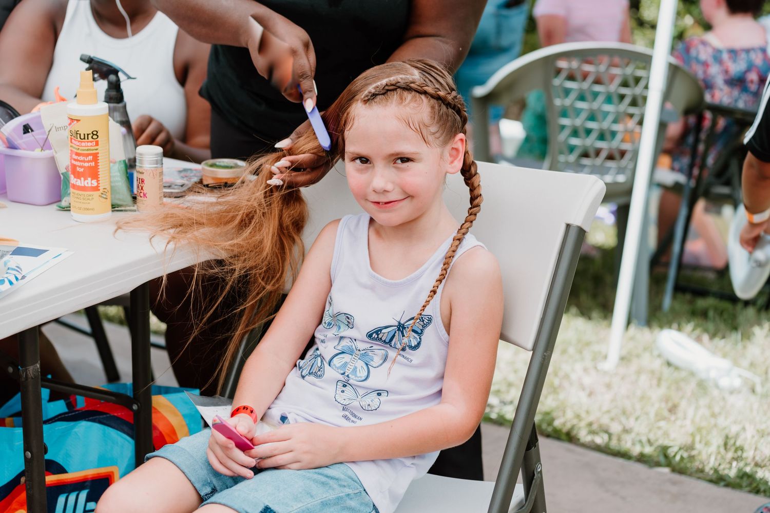 A person sits at an outdoor table while getting their hair styled at a community event.