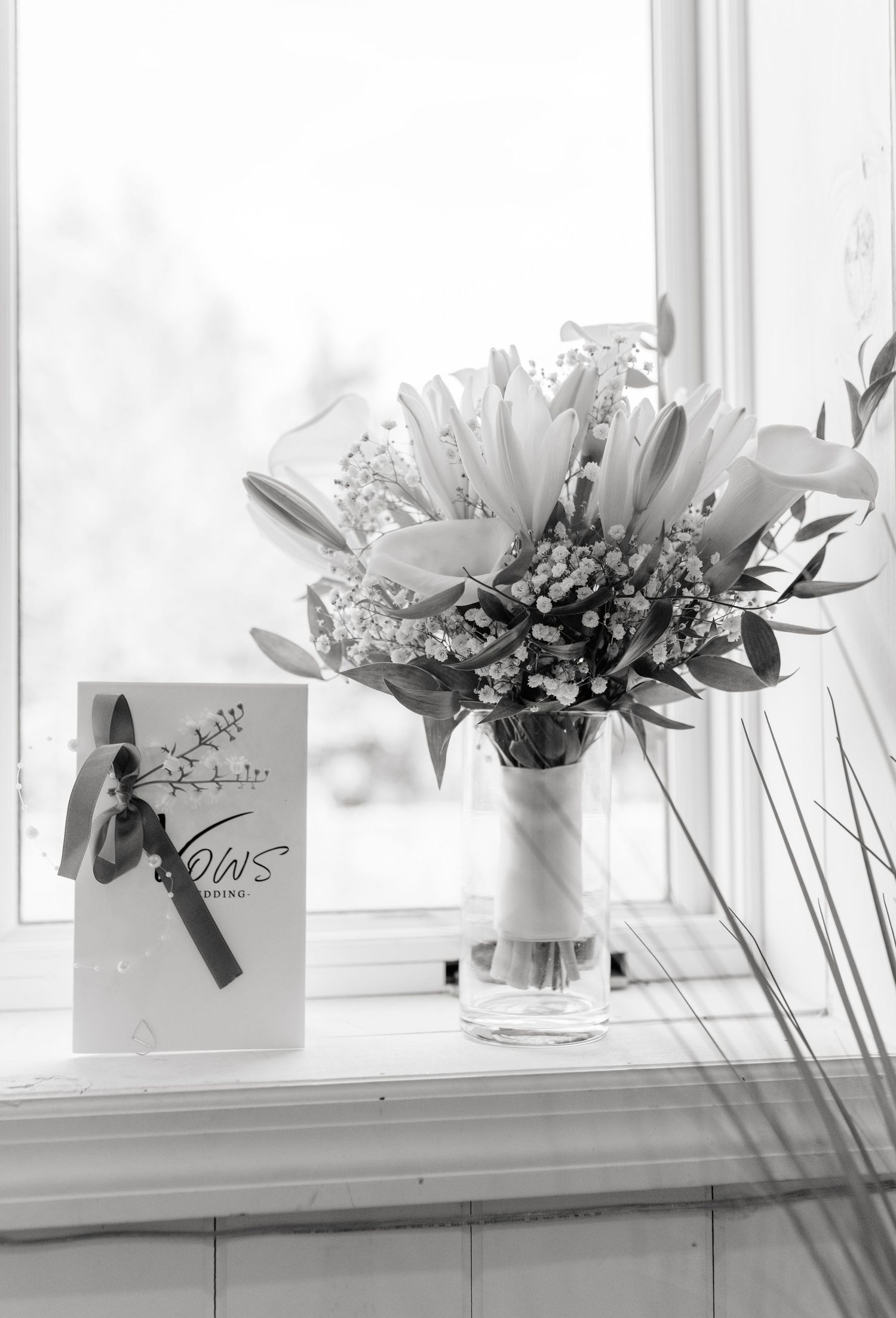 Black and white photo of a bridal bouquet of lilies in a glass vase beside a wedding vows booklet on a windowsill.