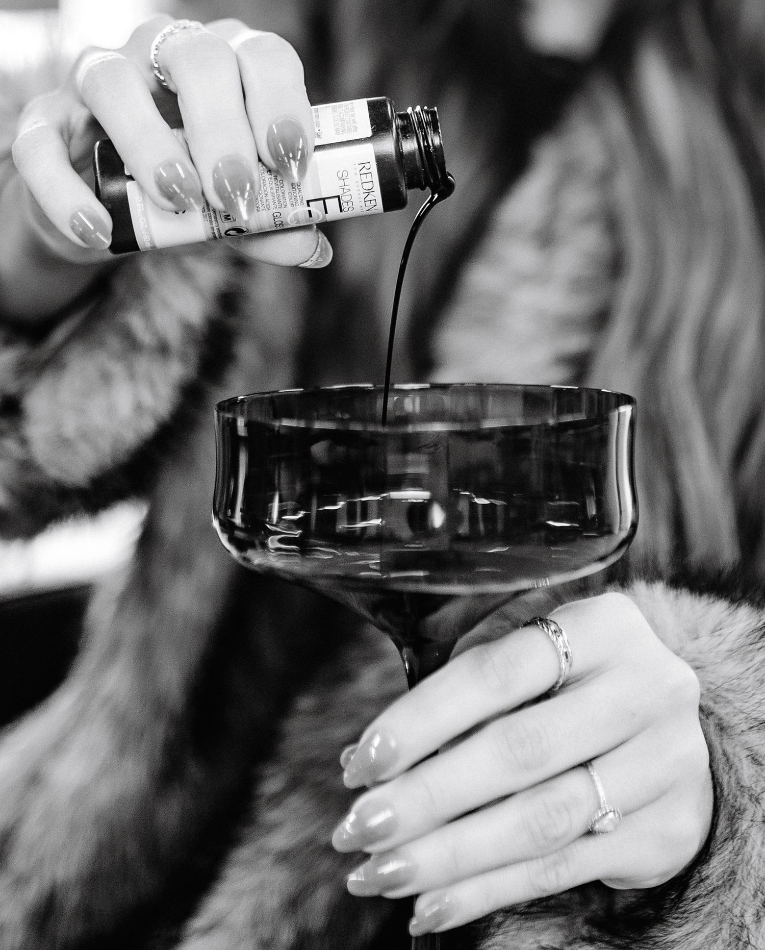 A black and white photo shows hands pouring a dark liquid into a cocktail glass from a small bottle.