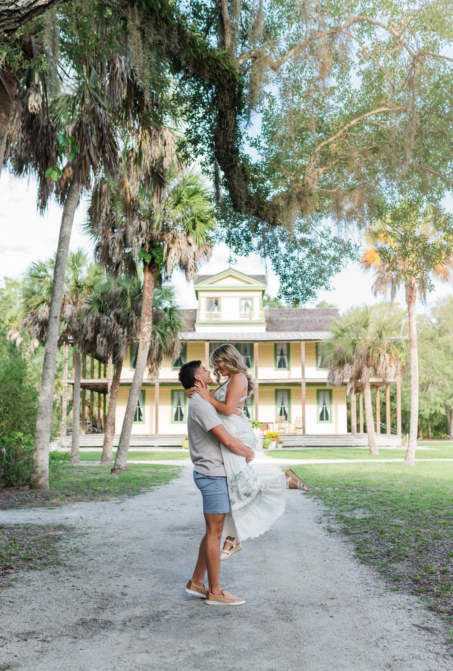 A couple shares a romantic lift embrace on a tree-lined path before a historic yellow Southern mansion.