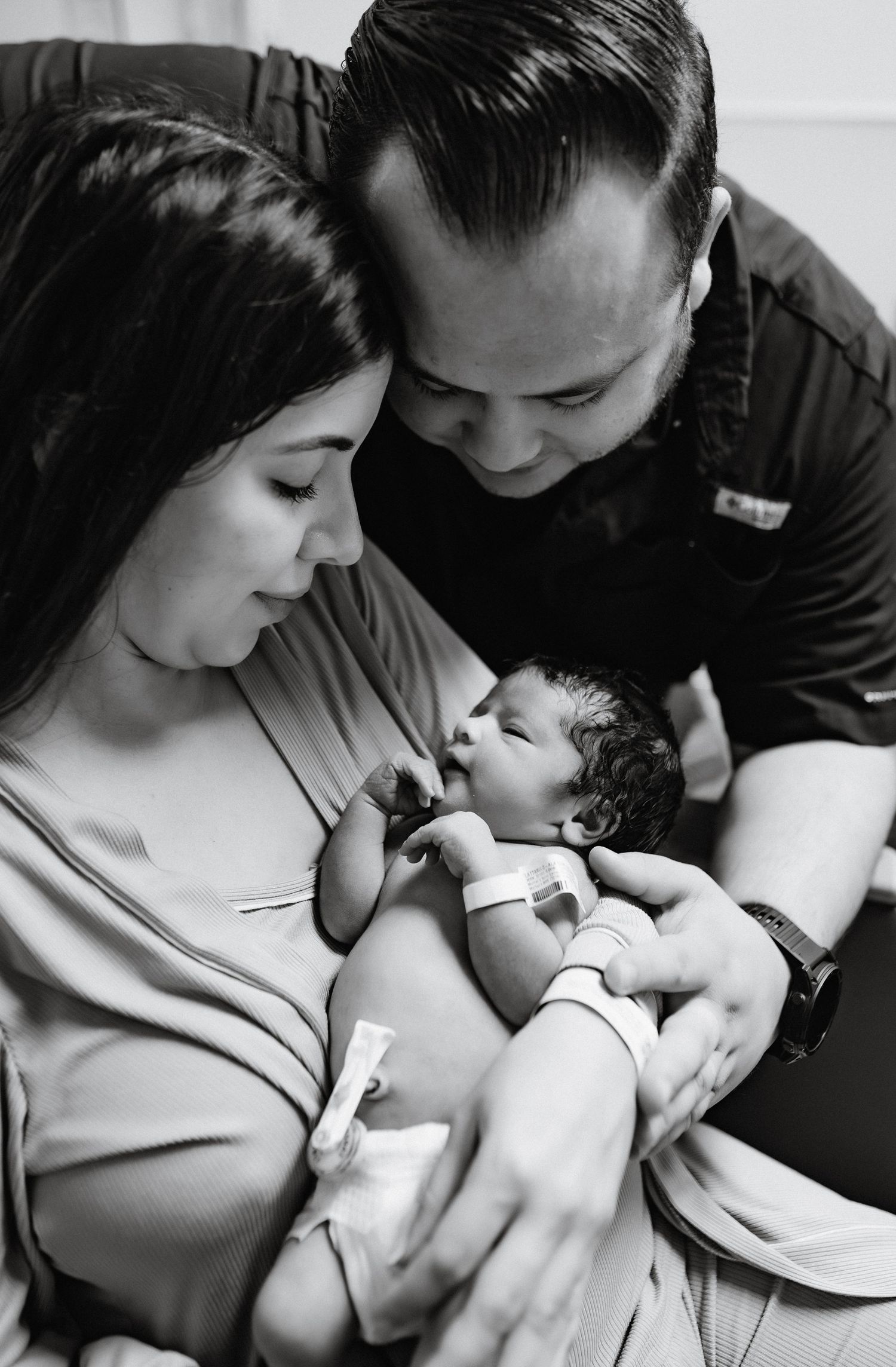 Black and white photo of new parents lovingly gazing at their newborn baby in a hospital room.