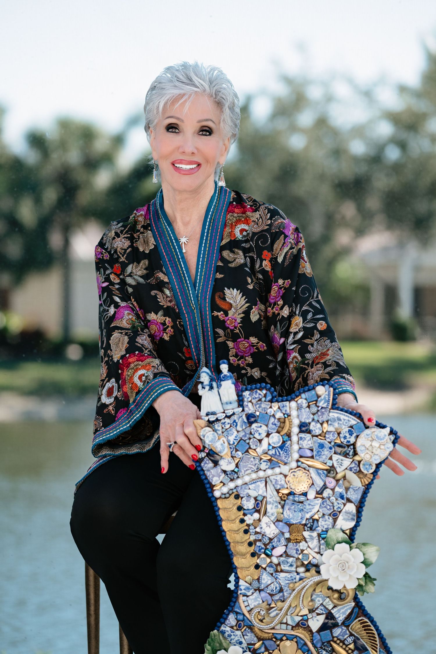 Woman in floral kimono poses with decorative blue and white mosaic heart sculpture outdoors near water.