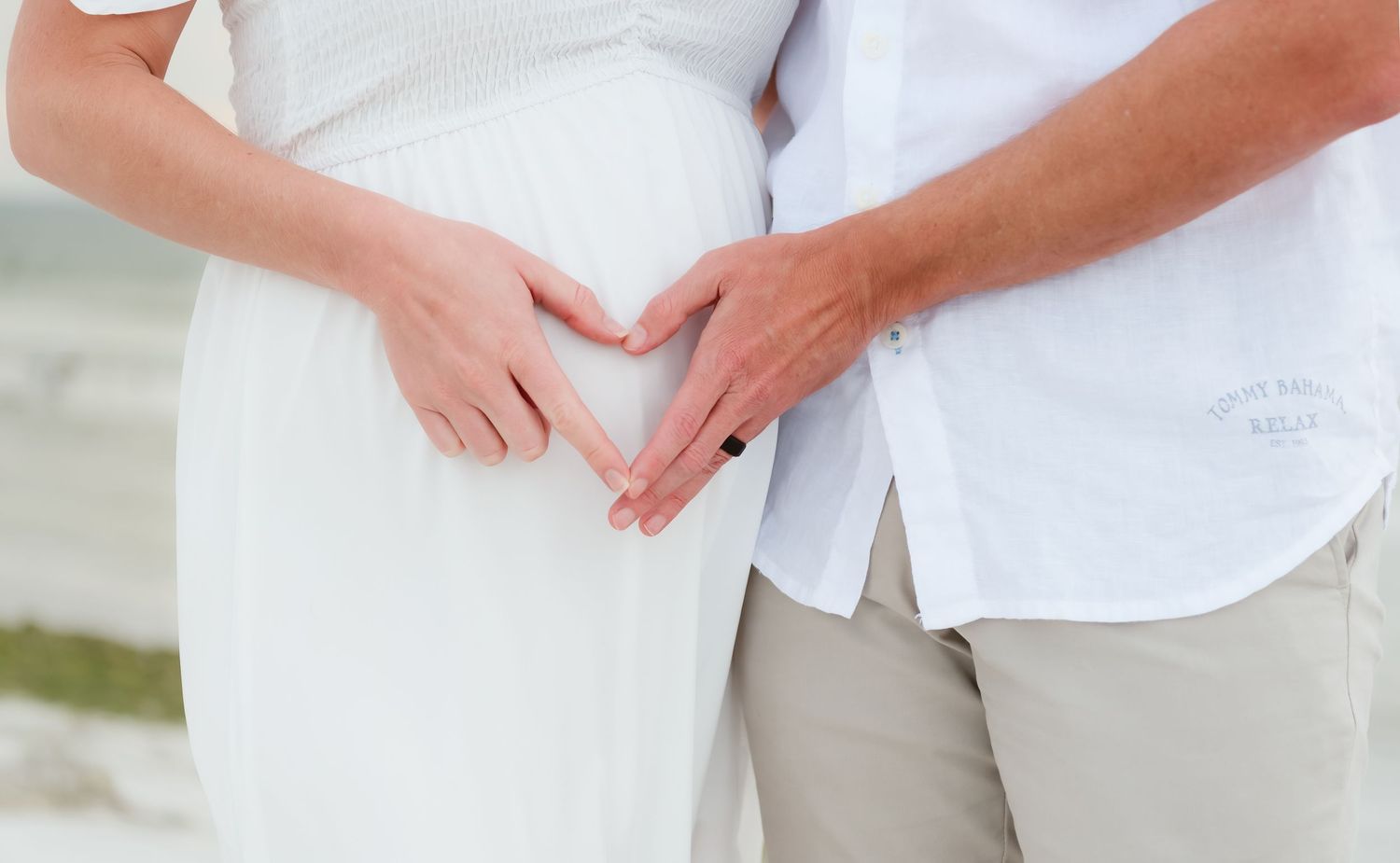 Couple making heart shape with hands on pregnant belly outdoors at beach in white and beige outfits.