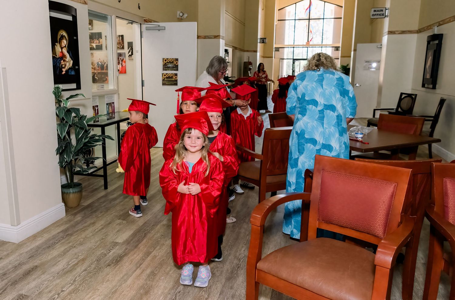 Children dressed in red and blue graduation caps and gowns lined up inside a room with leather chairs and gallery walls.