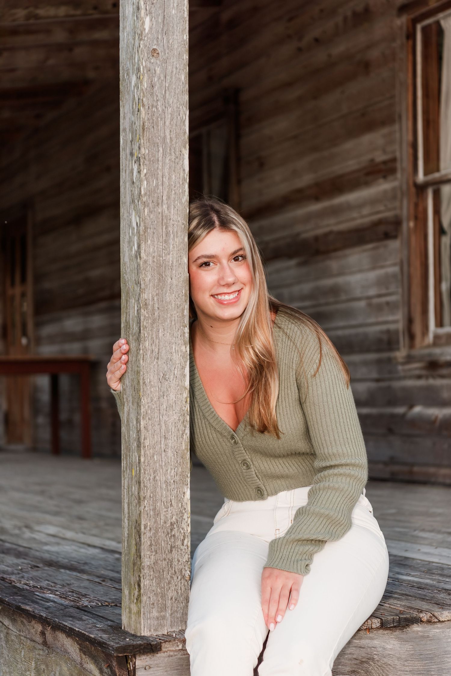 A person in a sage green sweater and white pants poses beside a rustic wooden post on an old porch.