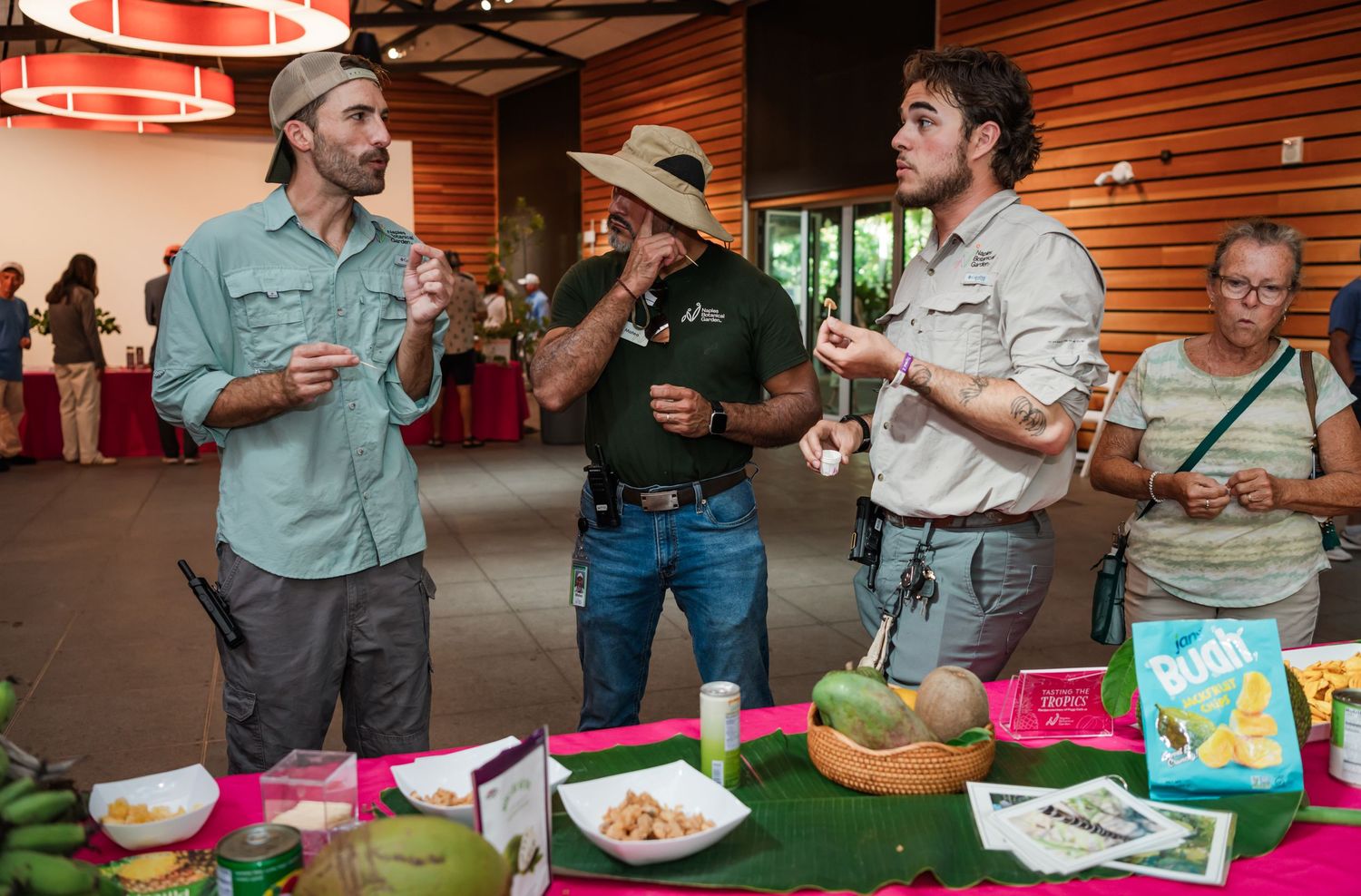 Three men in outdoor attire discuss tropical foods at a colorful display table during an indoor food event.