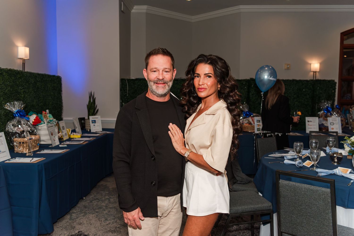 Two people pose together at an elegant event with blue-decorated tables and party decor in the background.