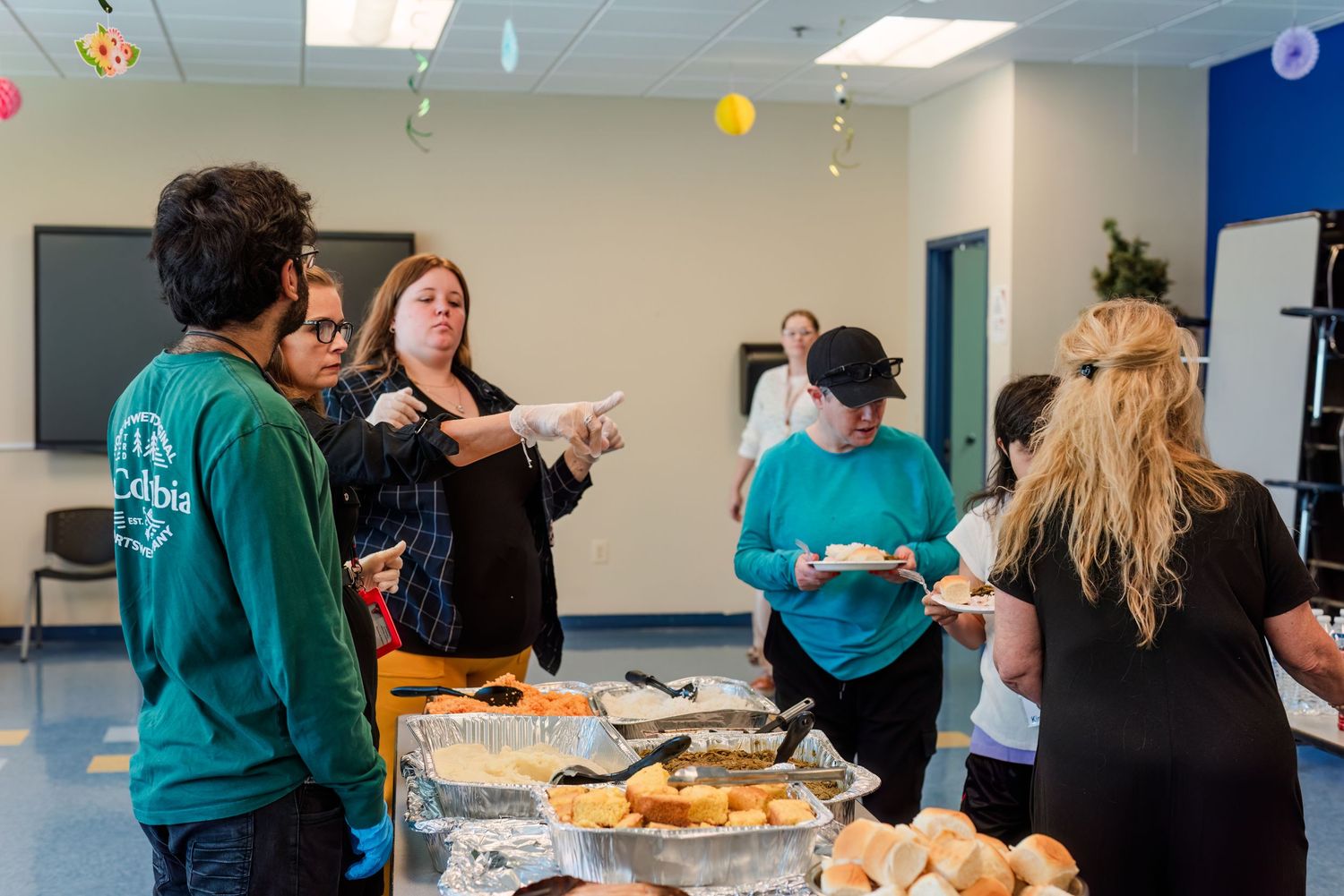 People gather around a buffet table with food during a casual indoor event wearing teal and black clothing.